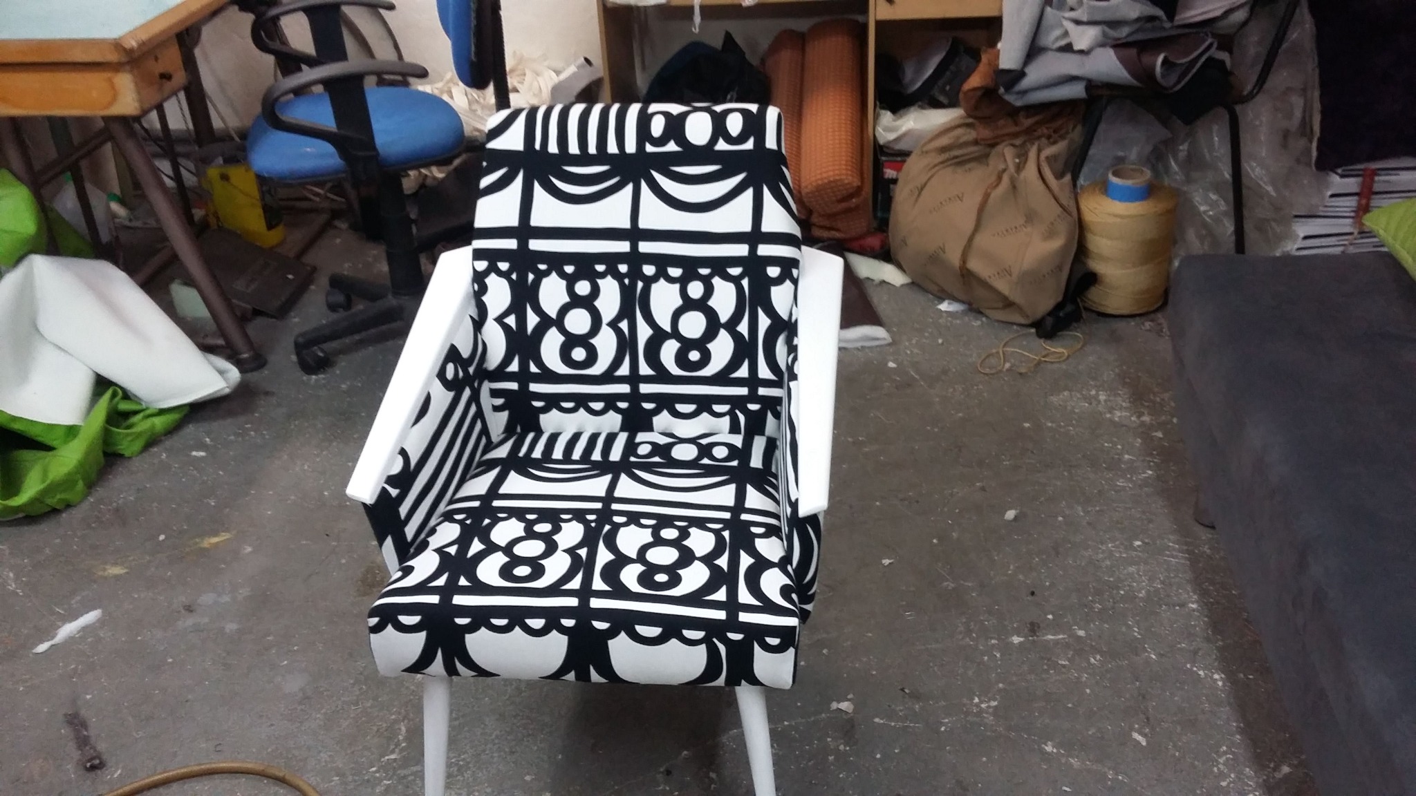 Odnowione krzesło z białymi ramami i tapicerką w czarno-białe geometryczne wzory, stojące w warsztacie z widocznymi narzędziami i materiałami.