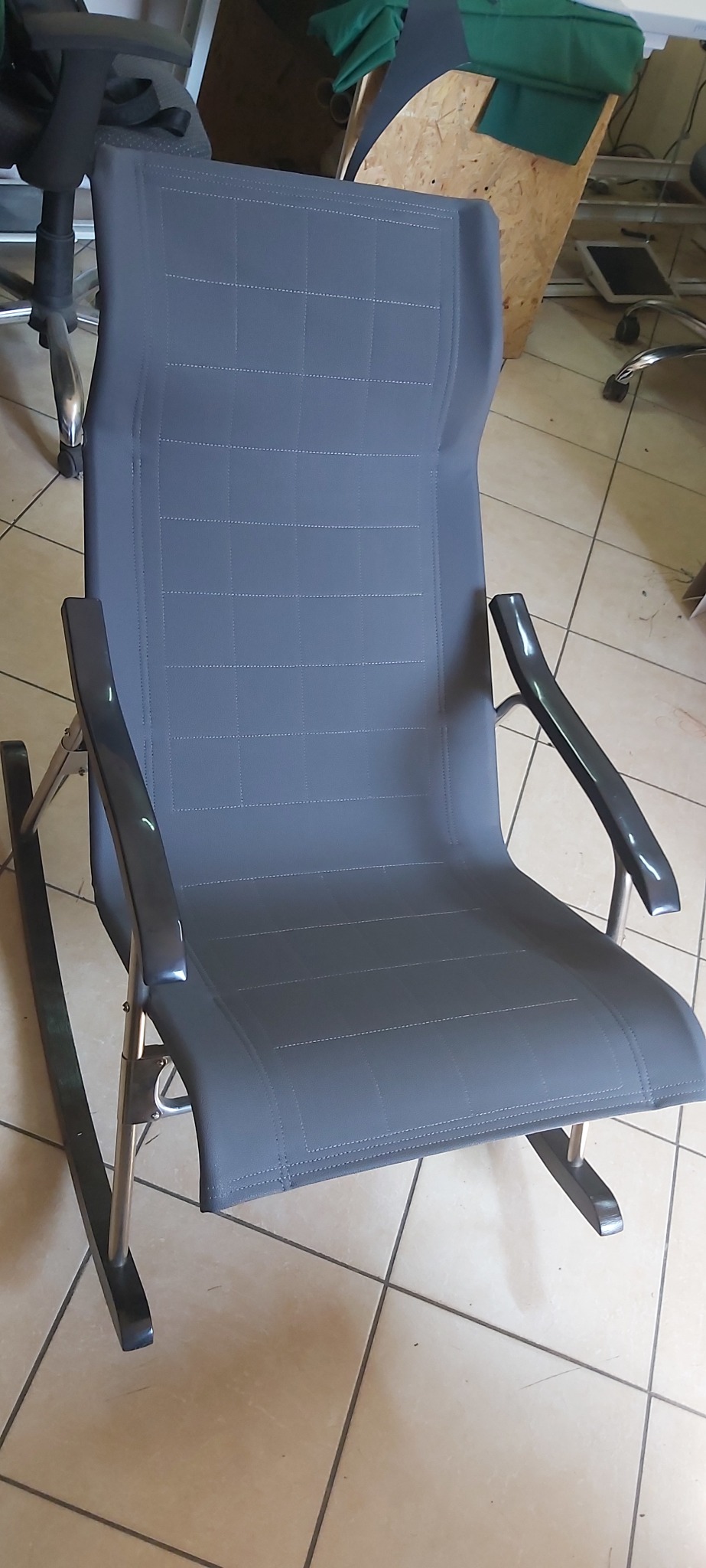 Szare krzesło bujane z tapicerowanym siedziskiem i oparciem, wykończone stebnowaniem, na metalowej ramie, widoczne w warsztacie.