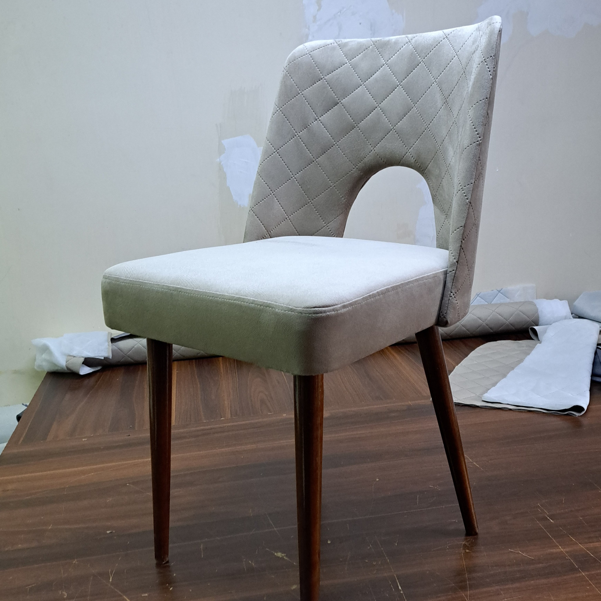 Beżowe krzesło z pikowanym oparciem i okrągłym wycięciem, stojące na drewnianym stole, obok leżą skrawki materiału.