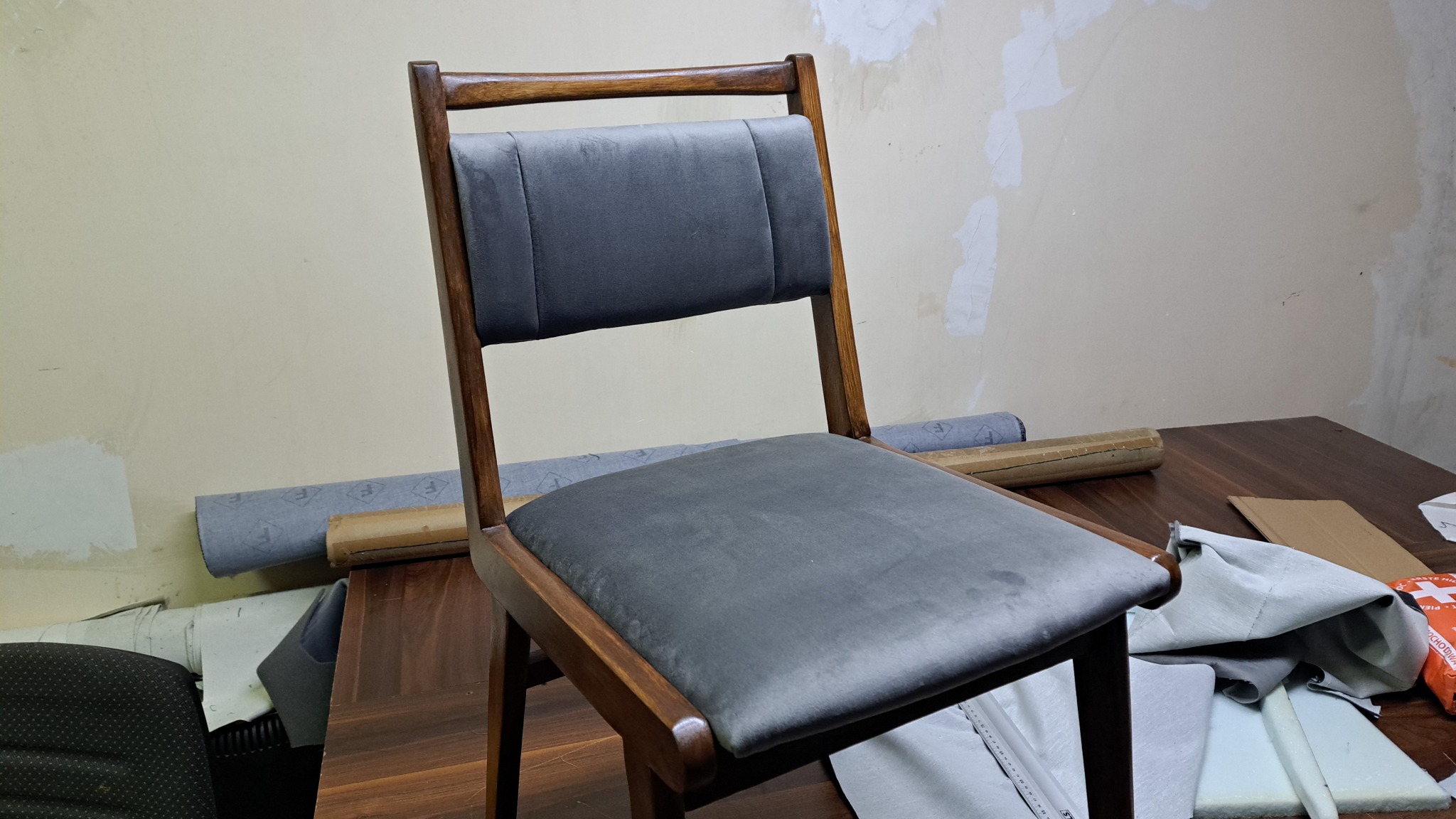 Drewniane krzesło z szarym, welurowym obiciem, widoczne materiały i narzędzia tapicerskie na stole w tle