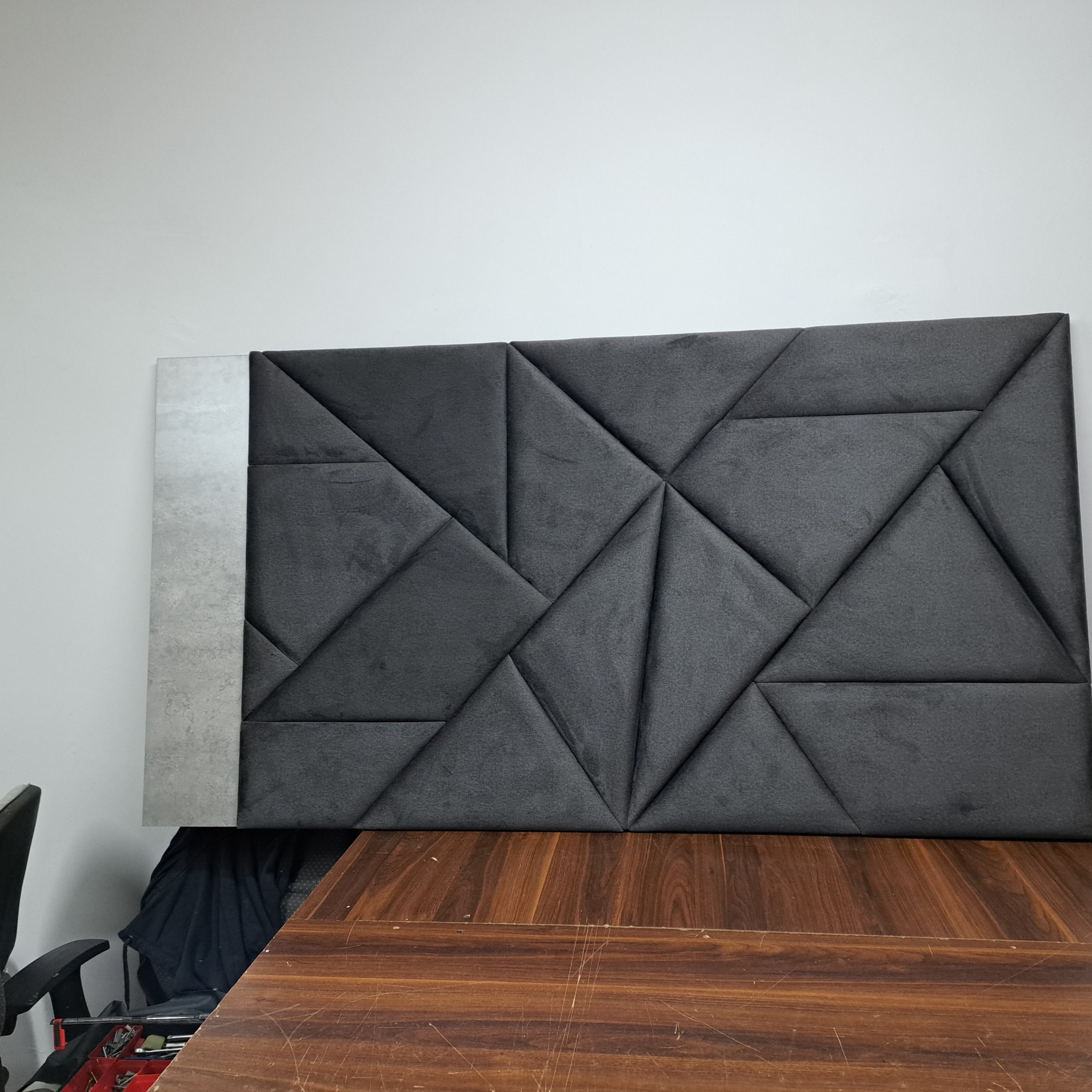 Panel ścienny z szarej tapicerki z geometrycznym wzorem trójkątów, z szarą płytą po lewej stronie, umieszczony nad drewnianym blatem.