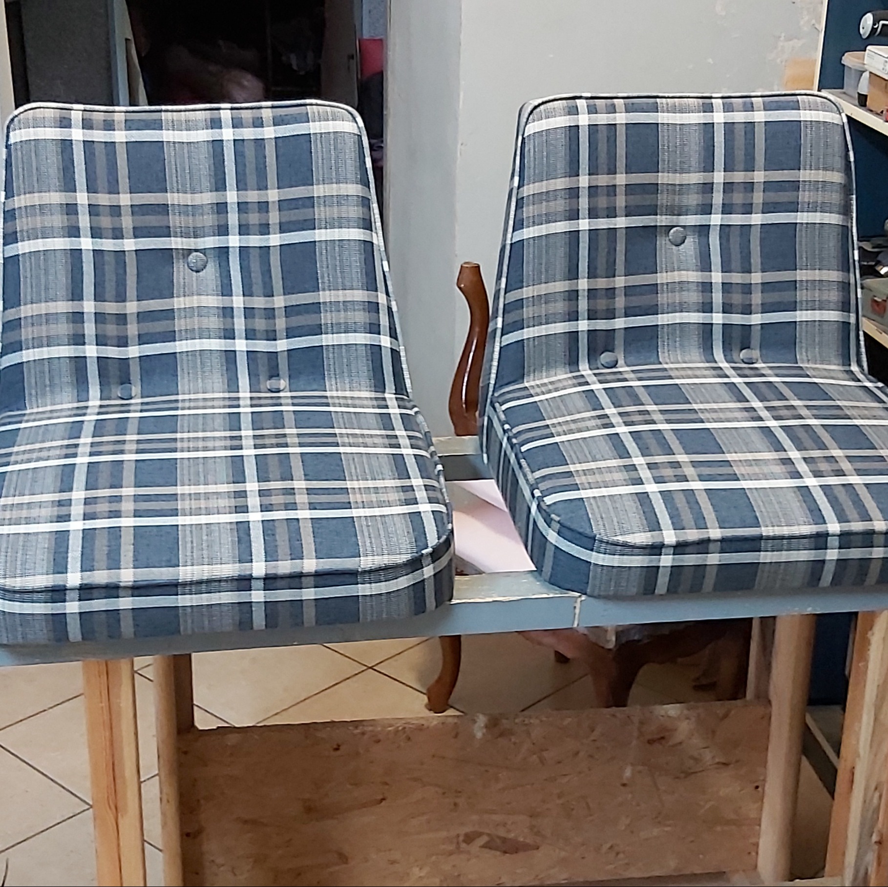Dwa krzesła po renowacji tapicerskiej, obite tkaniną w niebiesko-szarą kratę z guzikami, stojące na warsztatowym stole.