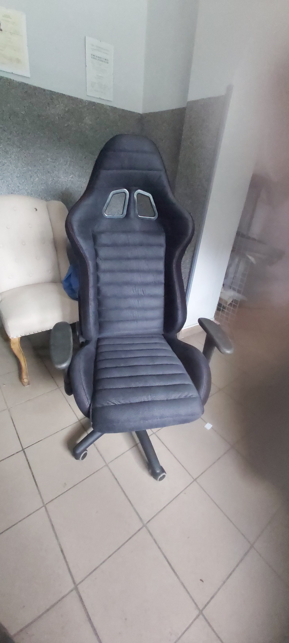 Czarno-szary fotel biurowy w stylu sportowym z widocznymi przeszyciami, stojący na kafelkach, z krzesłem obok i dokumentami na ścianie w tle.