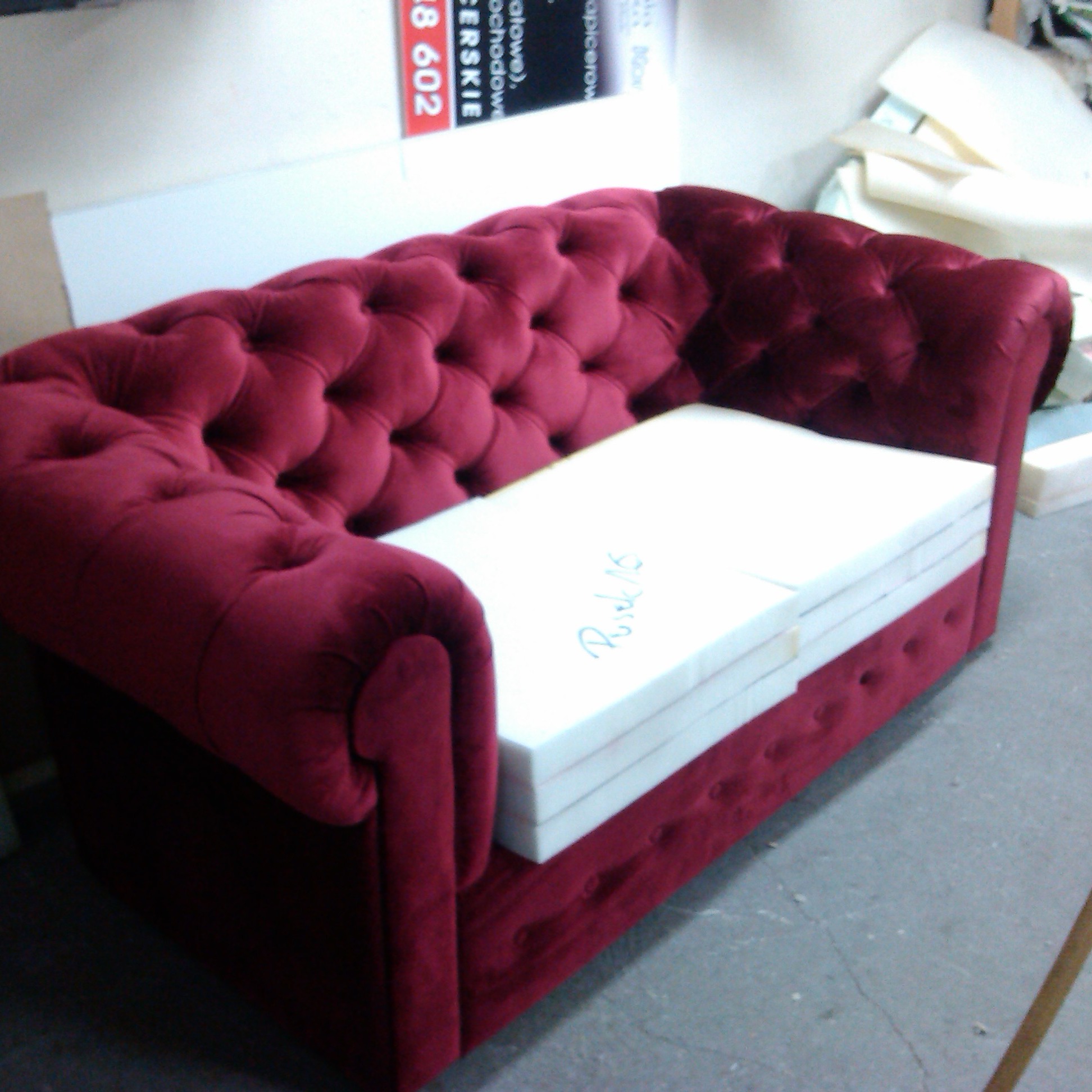 Czerwona sofa typu chesterfield w trakcie renowacji, z białymi piankowymi elementami siedziska ułożonymi na wierzchu, widoczne pikowania i guziki, w tle materiały i narzędzia warsztatowe.