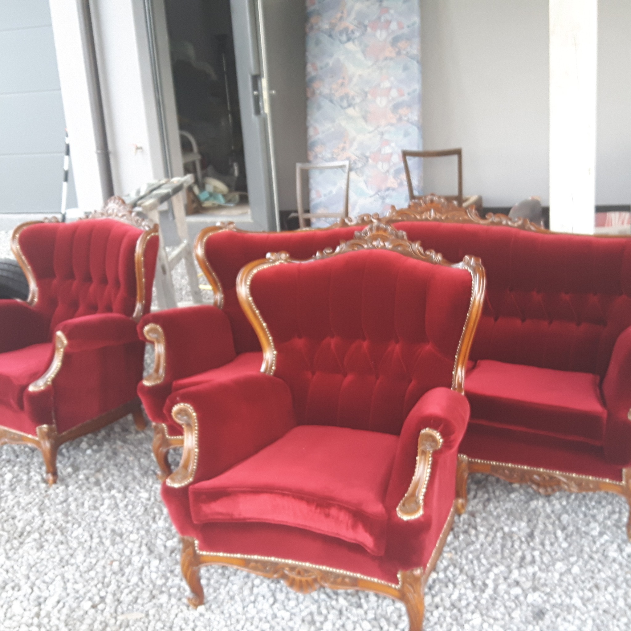 Komplet wypoczynkowy w stylu Ludwika XV z czerwonego weluru i rzeźbionego drewna, składający się z sofy i dwóch foteli typu uszak, ustawiony na żwirowym podłożu przed budynkiem z otwartymi drzwiami...