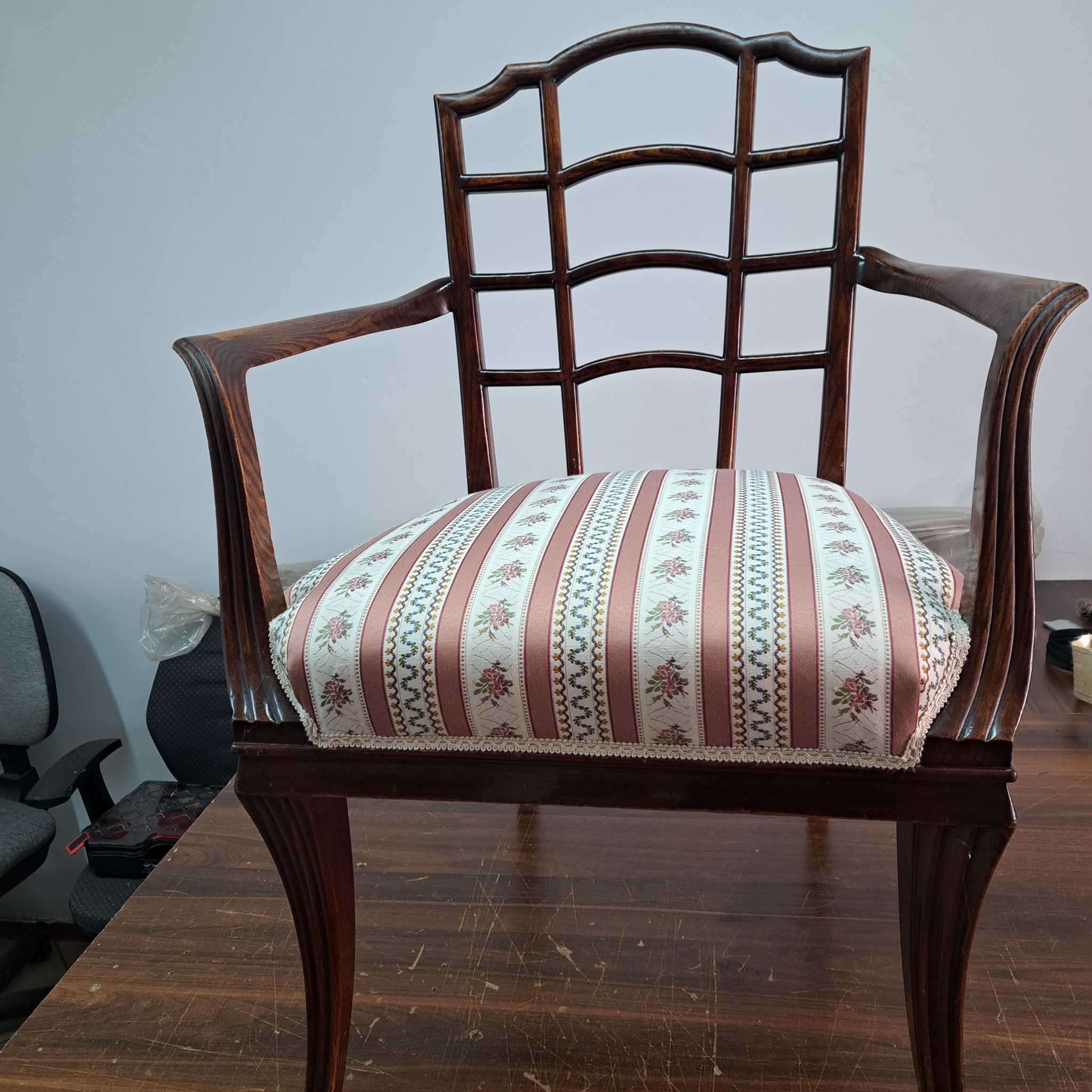 Odrestaurowane krzesło z ciemnego drewna z tapicerowanym siedziskiem w biało-różowe pasy z motywem kwiatowym, widoczne na tle biurowym z krzesłem obrotowym.