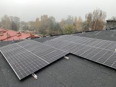 Panele fotowoltaiczne zamontowane na płaskim dachu pokrytym papą, widok z góry w pochmurny dzień. W tle budynki i drzewa.