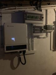 Biały falownik fotowoltaiczny zamontowany na szarej, pomalowanej ścianie w piwnicy, z widocznymi przewodami i skrzynkami elektrycznymi powyżej. Delikatna poświata na froncie urządzenia.