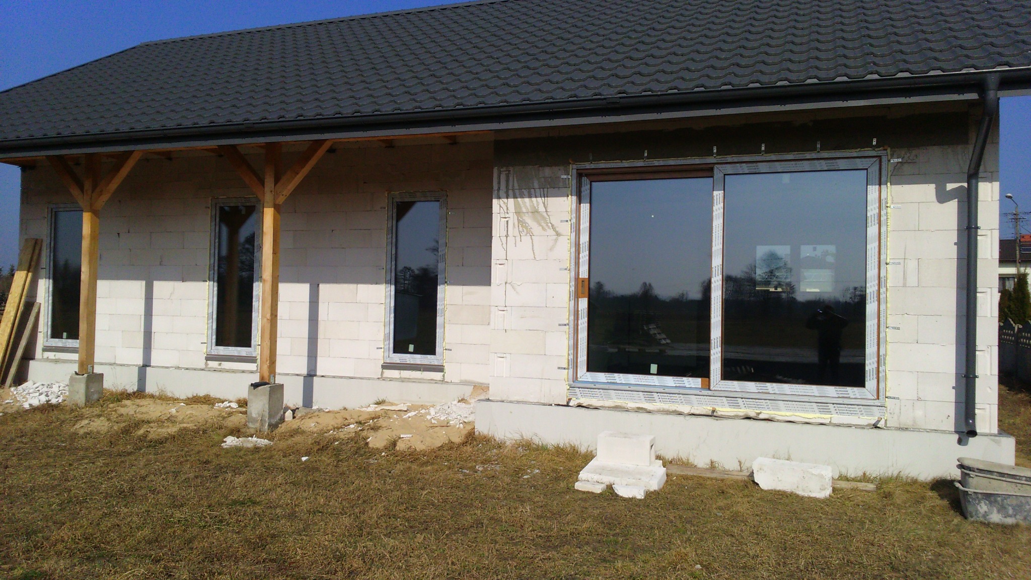 Dom w trakcie budowy z zamontowanymi oknami, widoczne białe bloczki, drewniane podpory ganku i przygotowane podłoże.