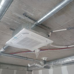 Krzysztof Pyc KLIMA-HVAC - Kasetonowa jednostka klimatyzacji pod sufitem w folii ochronnej, widoczne rury wentylacyjne i instalacje. Surowy betonowy sufit i ściany w trakcie wykończenia.