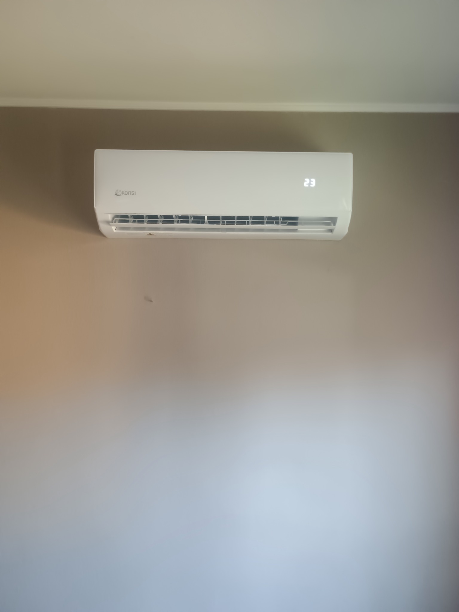 Biała klimatyzacja marki Konsi na beżowej ścianie. Na urządzeniu widoczny wyświetlacz z temperaturą 23 stopnie.