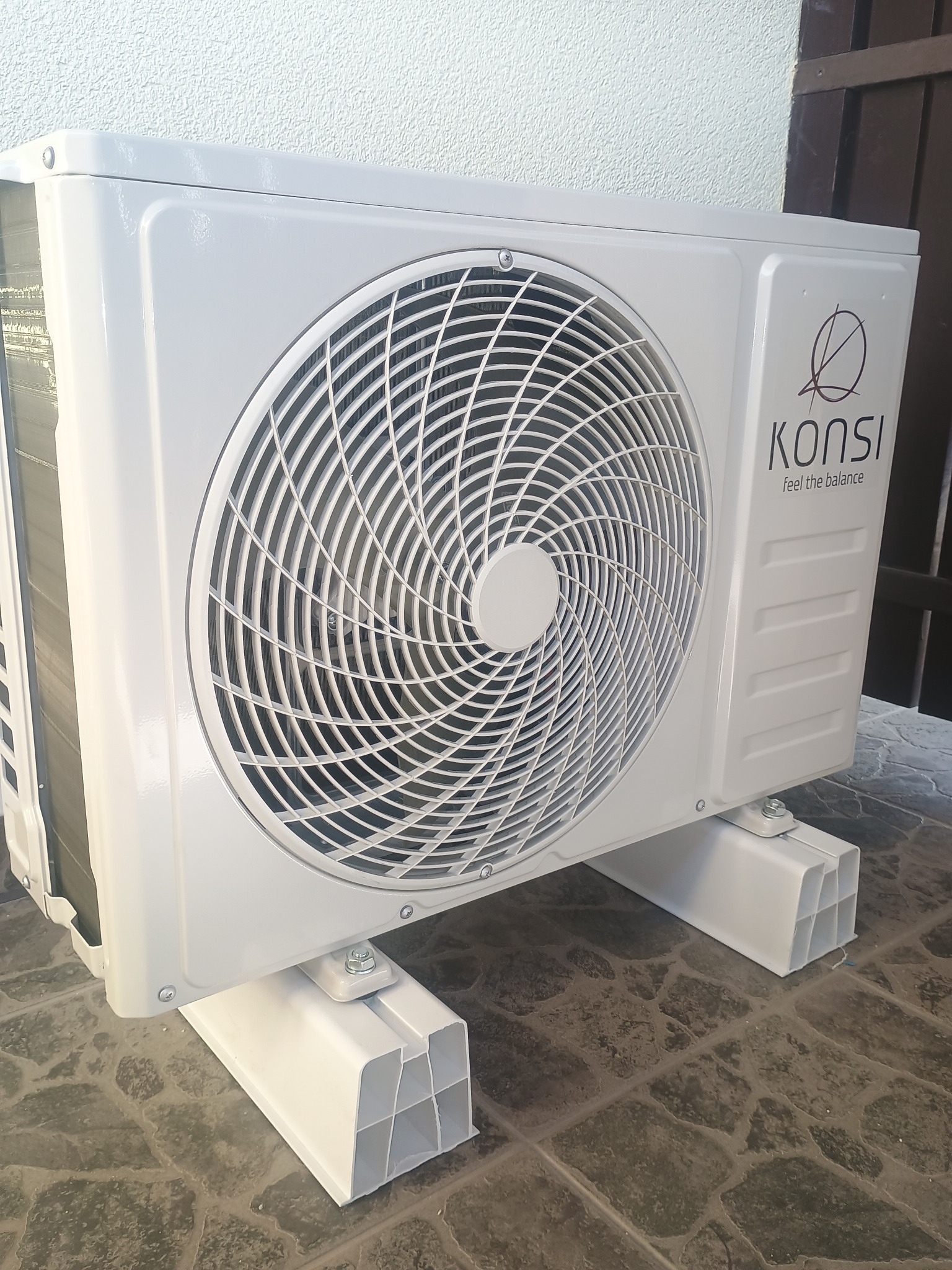 Biała jednostka zewnętrzna klimatyzacji marki Konsi, zamontowana na białych wspornikach na zewnątrz budynku, z widocznym logo i napisem 'feel the balance'.