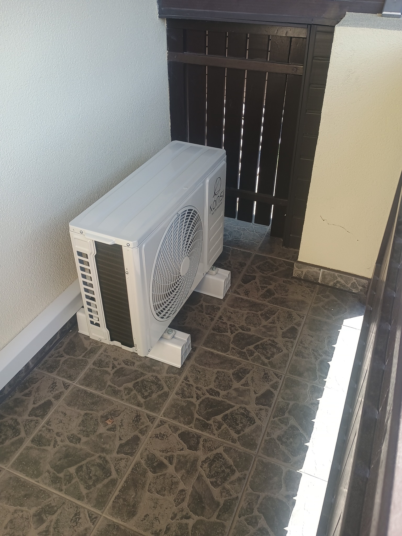 Biały klimatyzator marki Konsi umieszczony na balkonie z ciemnymi płytkami, tuż obok drewnianej, ciemnej bramki.