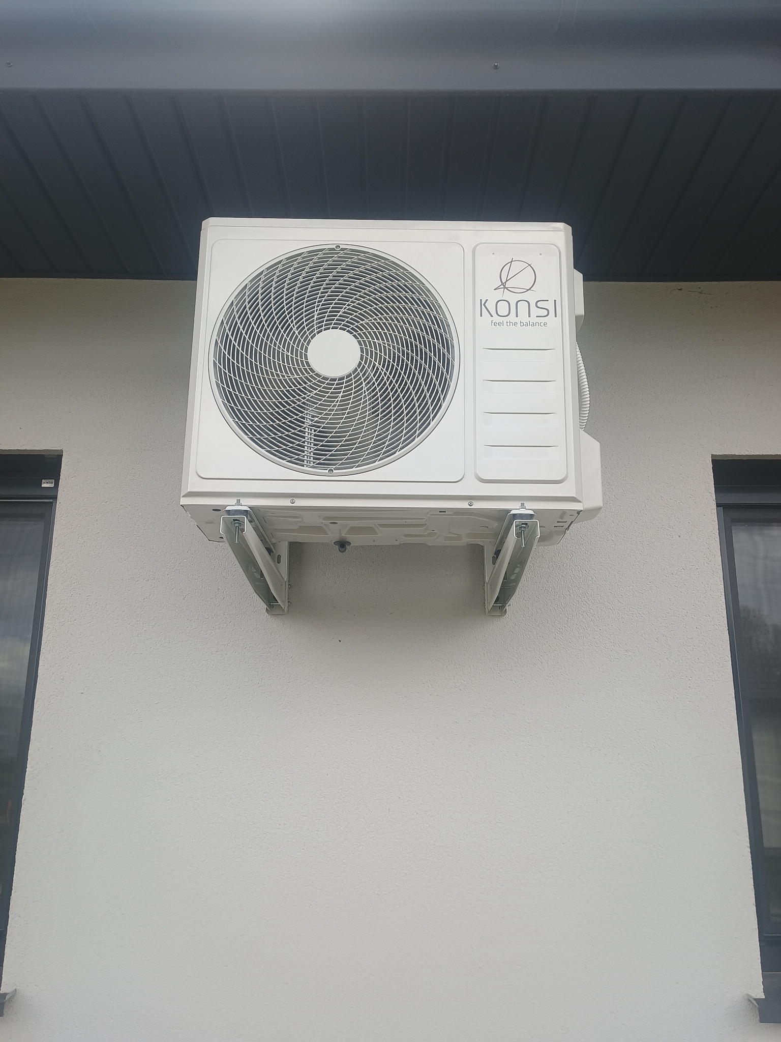 Biała jednostka zewnętrzna klimatyzacji marki Konsi, zamontowana na wspornikach na jasnej elewacji budynku. Widoczne okna i fragment podbitki dachowej.