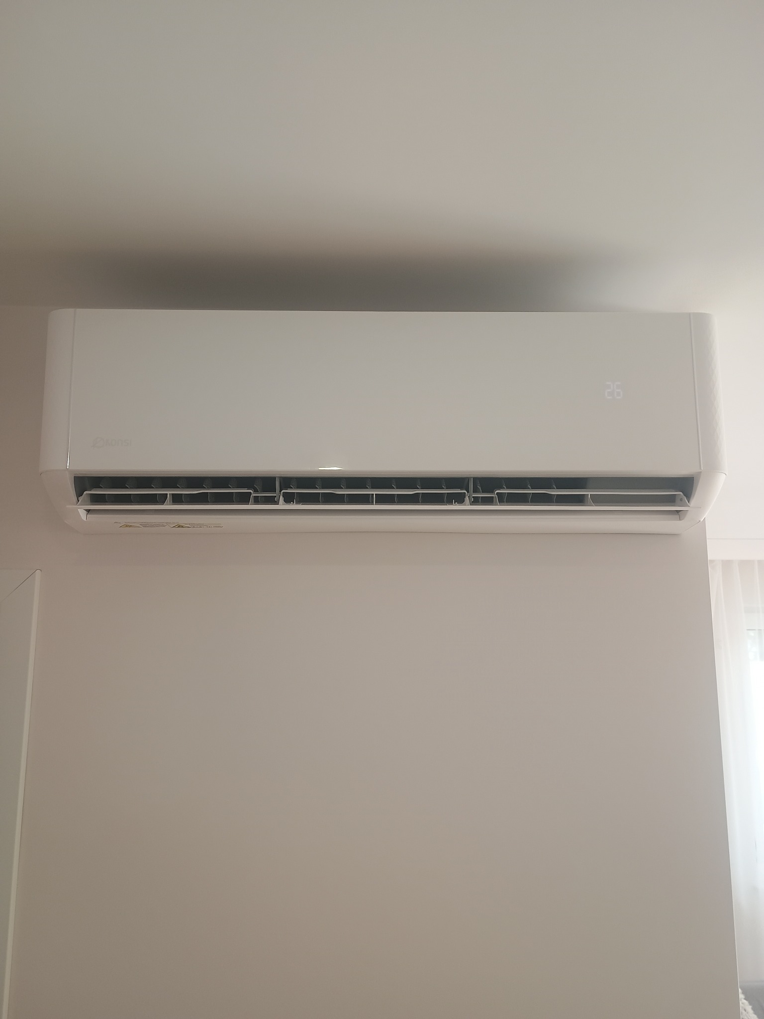 Biała klimatyzacja marki Konsi zamontowana na jasnej ścianie. Widoczny wyświetlacz z temperaturą 26 stopni. Minimalistyczny design, wnętrze mieszkalne.
