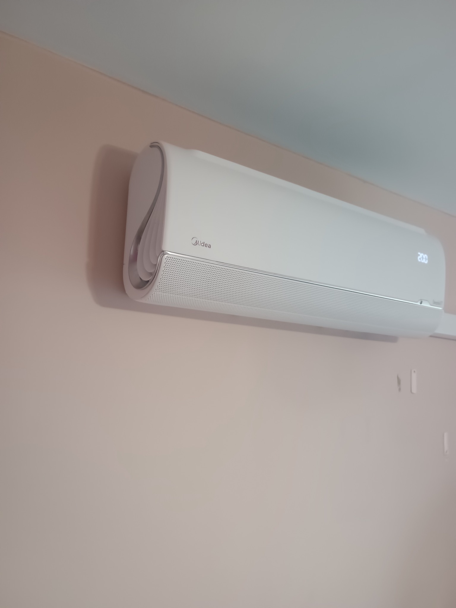 Nowoczesny, biały klimatyzator Midea zamontowany na pastelowej ścianie. Widoczne detale wykończenia i wyświetlacz temperatury. Minimalistyczny design, estetyczny montaż.