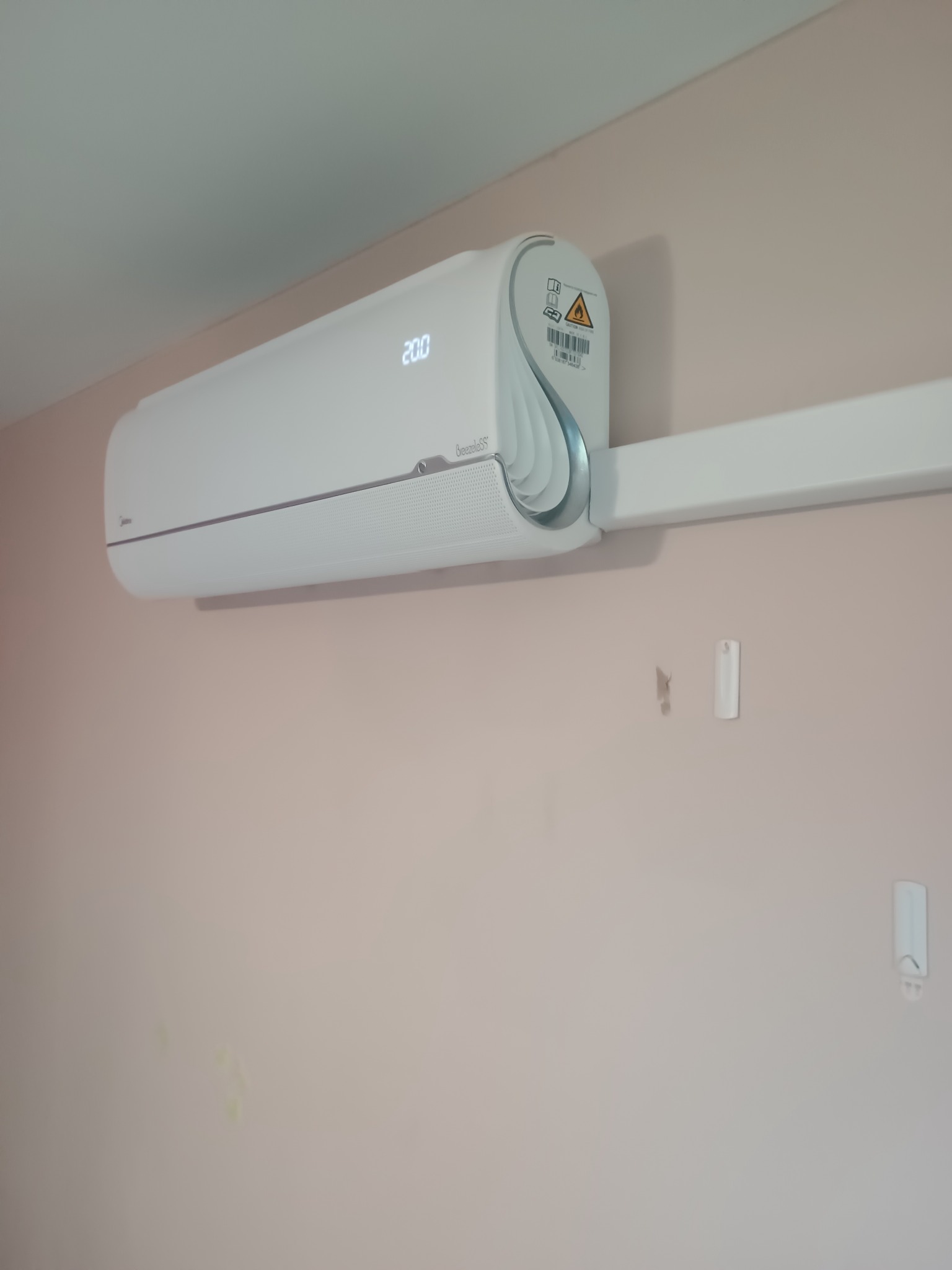 Biały klimatyzator Midea BreezeleSS zamontowany na ścianie w jasnym pomieszczeniu. Widoczny wyświetlacz z temperaturą 20 stopni Celsjusza.
