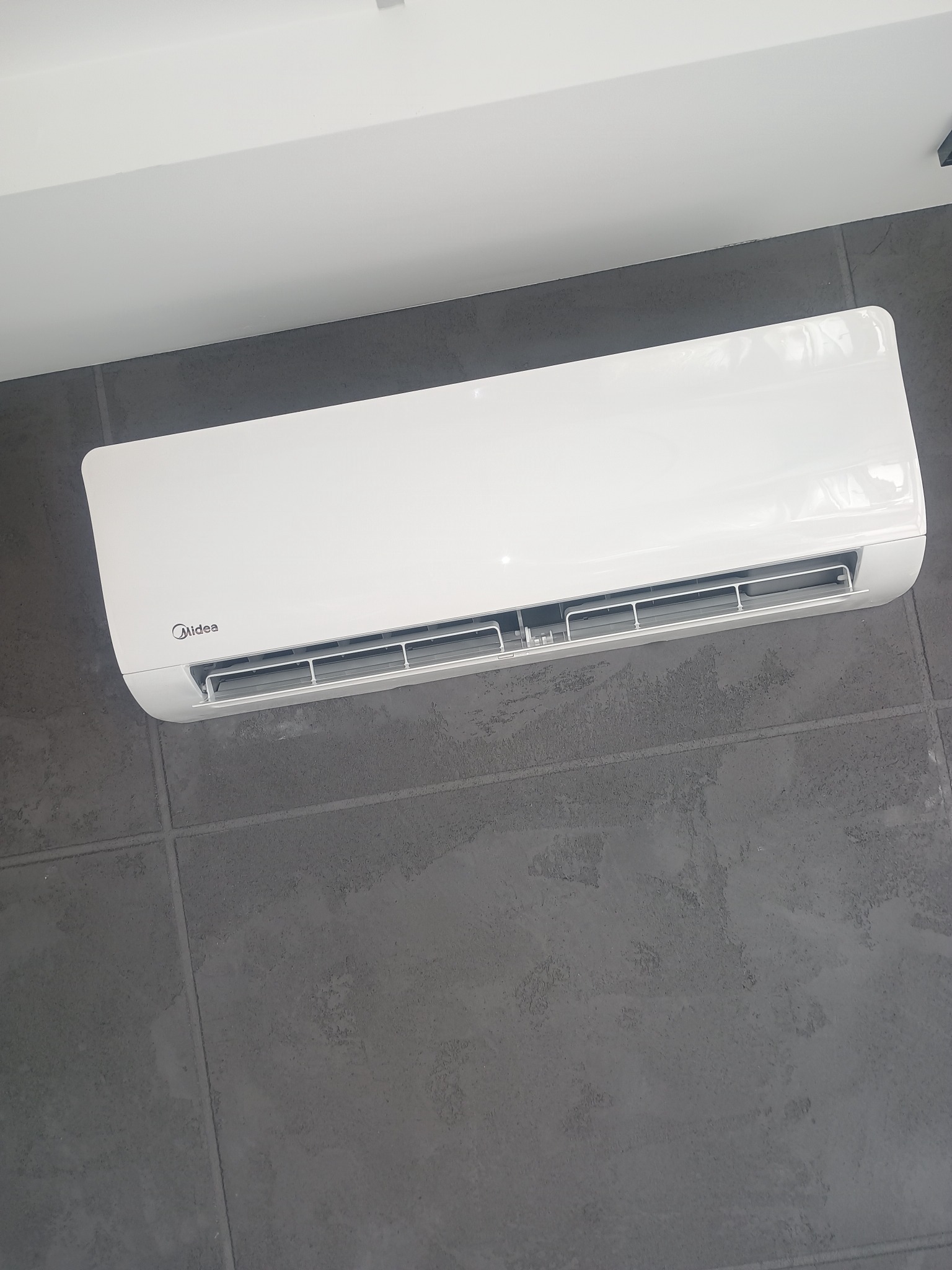 Biała klimatyzacja Midea na szarej ścianie z płytek, widok z dołu. Minimalistyczny design, nowoczesne wnętrze,  oświetlenie sufitowe.