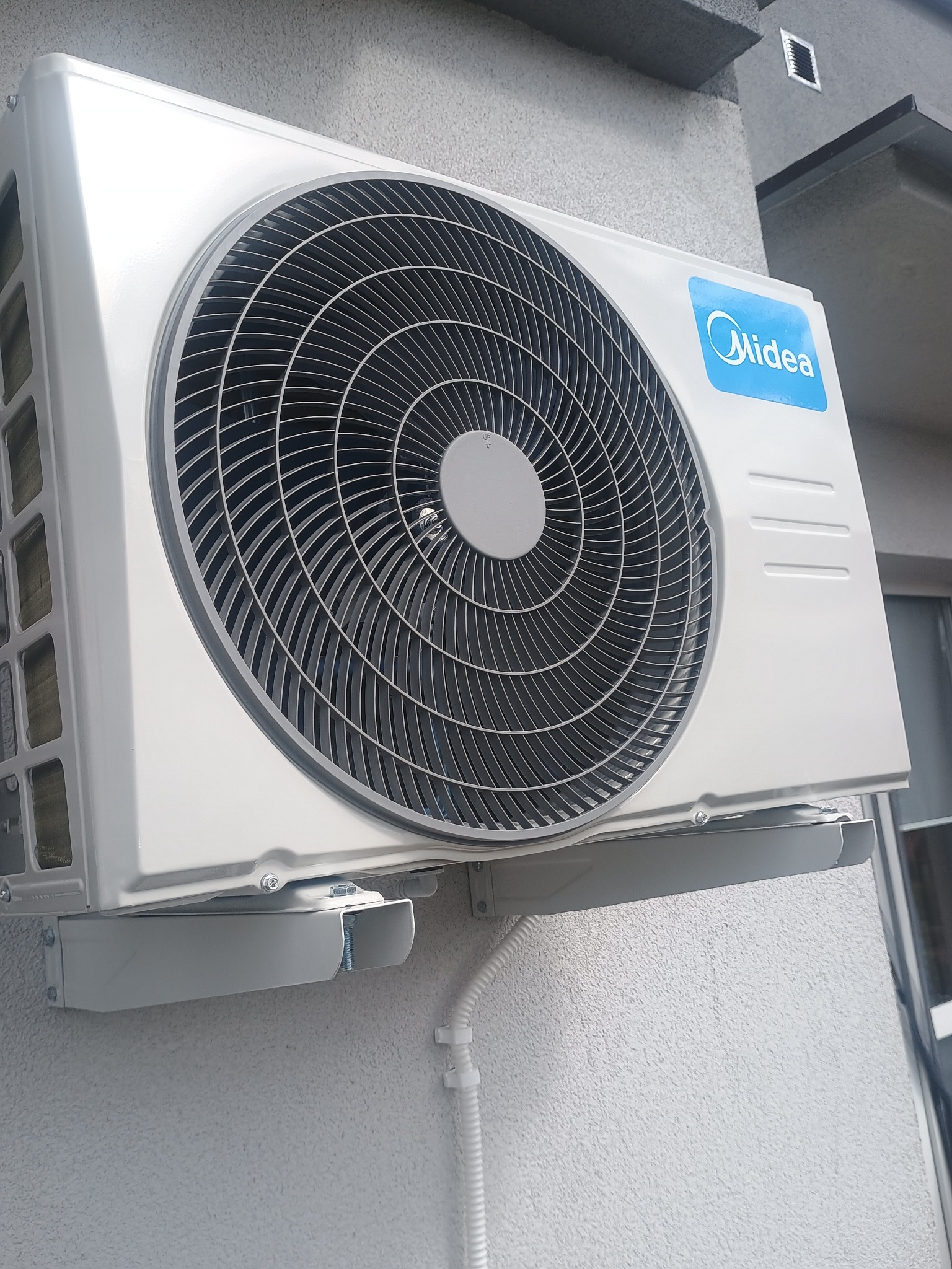 Zamontowany na zewnątrz, biały klimatyzator Midea z widoczną kratką wentylatora i przyłączami, na tle szarej ściany budynku.