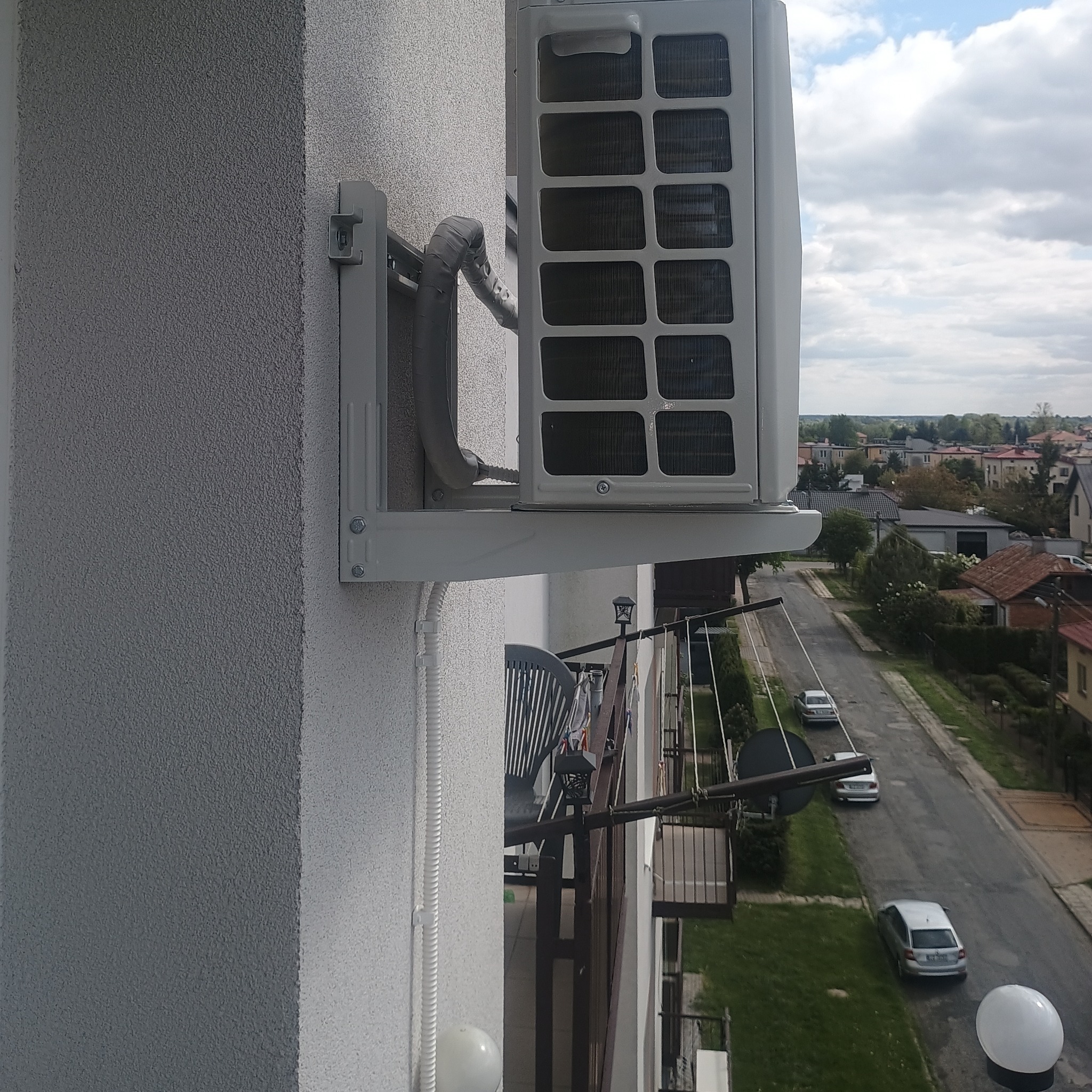 Zamontowany klimatyzator na balkonie budynku mieszkalnego, widok z boku. Na zewnątrz widoczna zabudowa miejska i pochmurne niebo. Elementy instalacji estetycznie ukryte.
