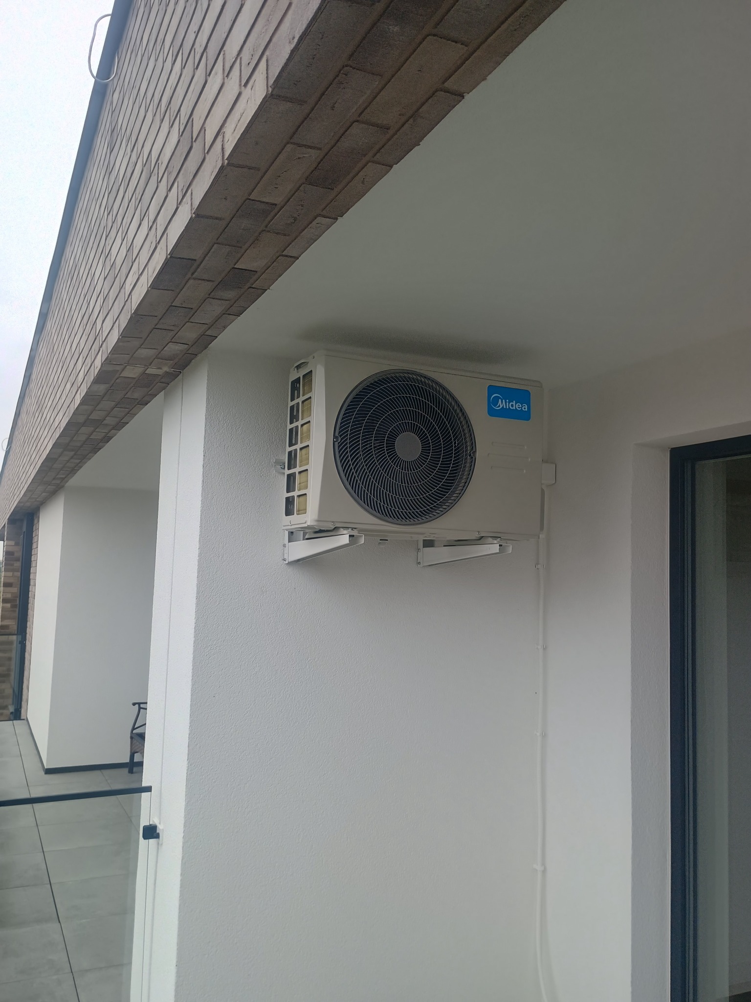 Zamontowana jednostka zewnętrzna klimatyzacji Midea na jasnej elewacji budynku z ceglanym wykończeniem. Widoczne przewody instalacyjne.