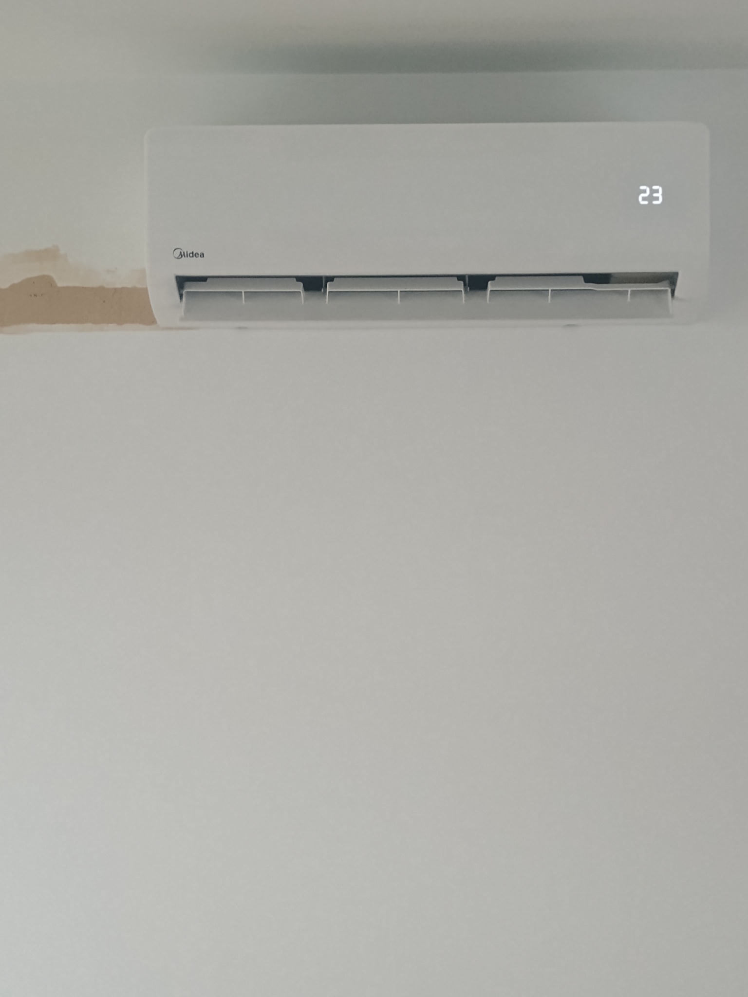 Biały klimatyzator Midea na ścianie z widocznymi śladami naprawy. Wyświetlacz pokazuje 23 stopnie Celsjusza. Ujęcie z dołu, centralne.