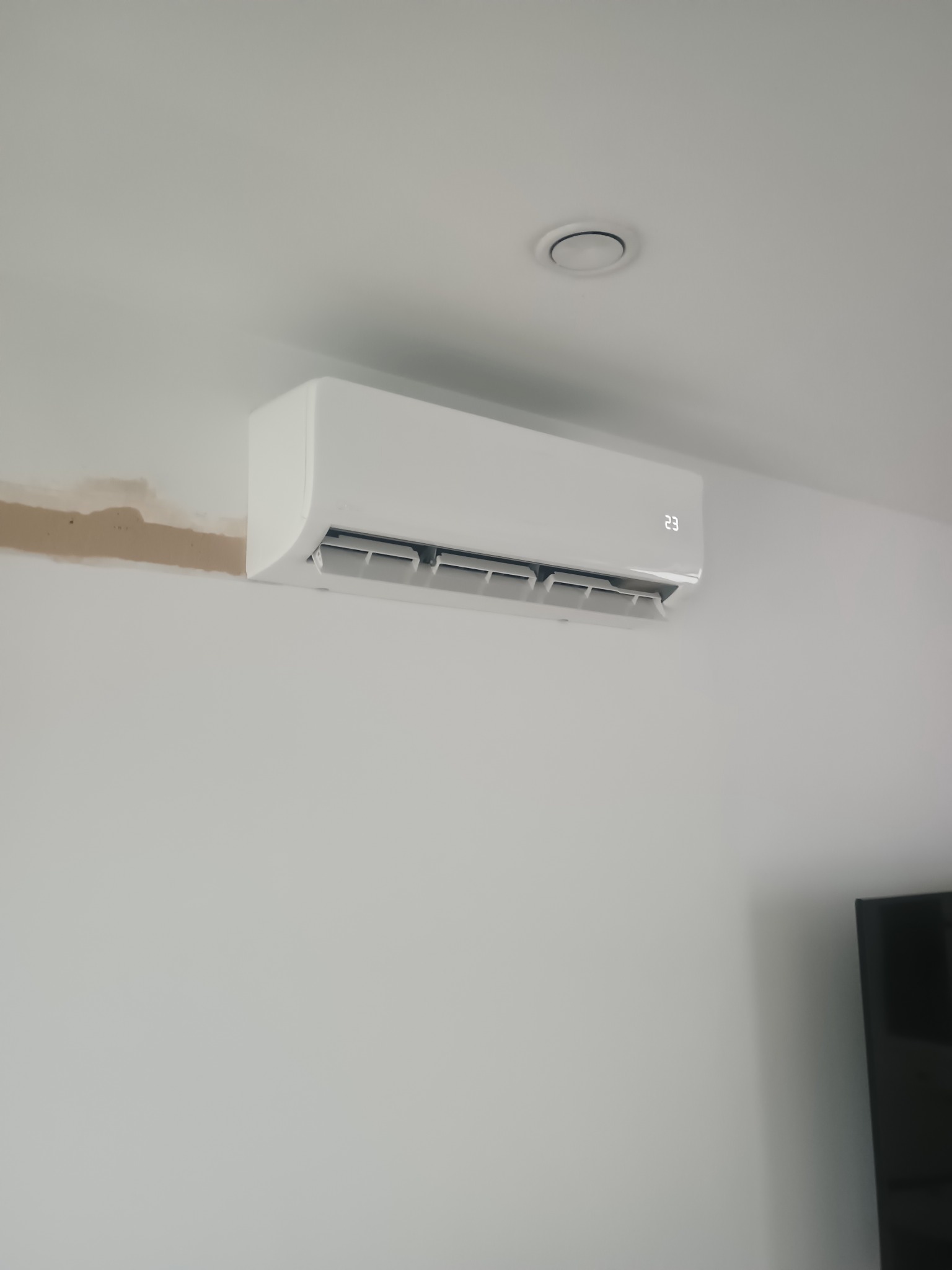 Biały klimatyzator ścienny z wyświetlaczem temperatury (23°C), zamontowany w jasnym wnętrzu, widoczne ślady po montażu na ścianie i element sufitu.