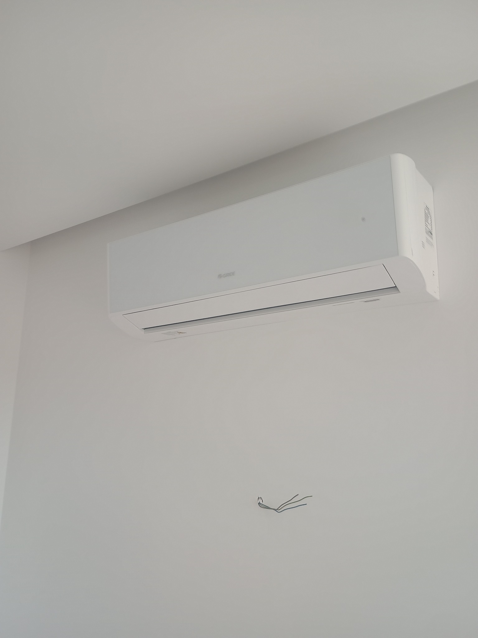 Biały klimatyzator marki Gree zamontowany na ścianie w jasnym pomieszczeniu. Widoczne wystające kable. Minimalistyczny design.