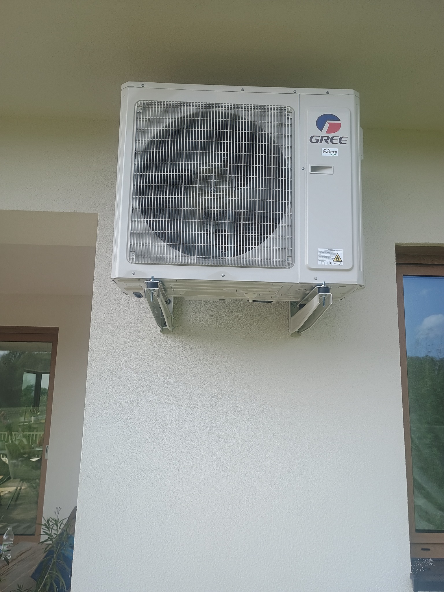Zamontowana na zewnątrz, biała jednostka klimatyzacji marki Gree, przymocowana do ściany budynku za pomocą metalowych wsporników. Widoczne okno i fragment rośliny.
