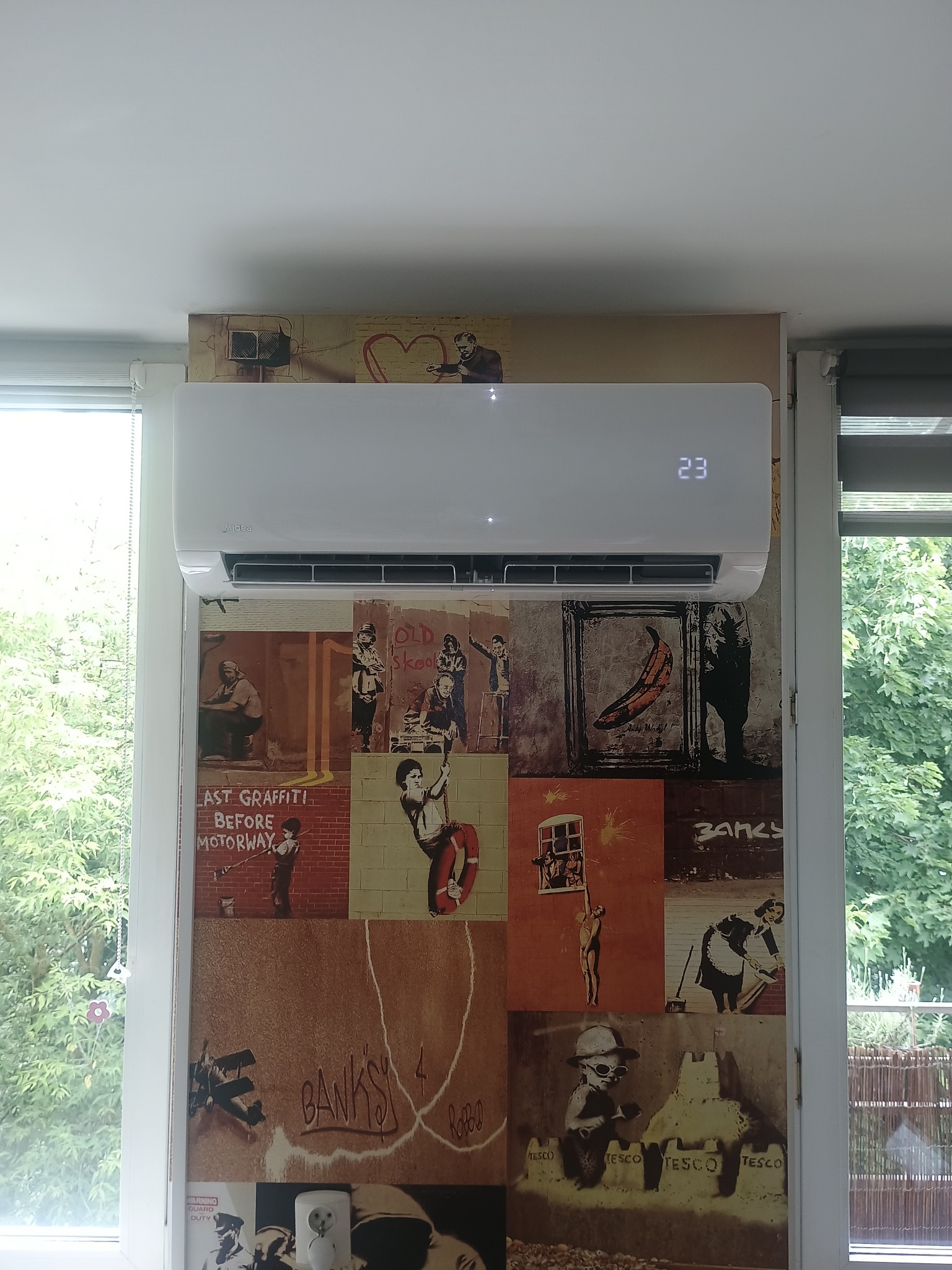 Biała klimatyzacja Midea na ścianie z grafikami Banksy'ego, ustawiona między oknami z widokiem na zieleń. Na wyświetlaczu klimatyzacji widoczna temperatura 23 stopnie.