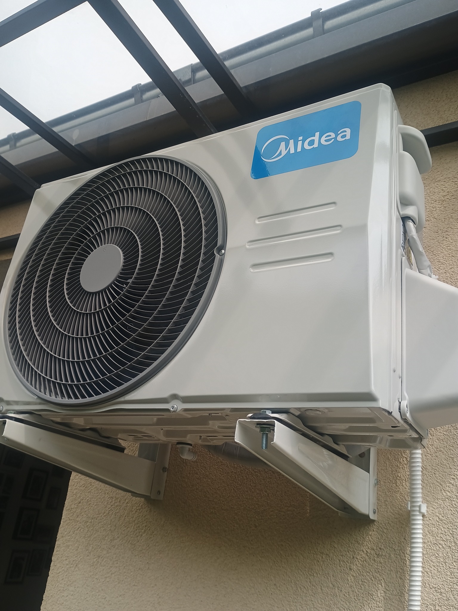 Biała jednostka zewnętrzna klimatyzacji Midea zamontowana na ścianie budynku, widoczna z dołu, z elementami instalacji i osłoną przeciwsłoneczną.