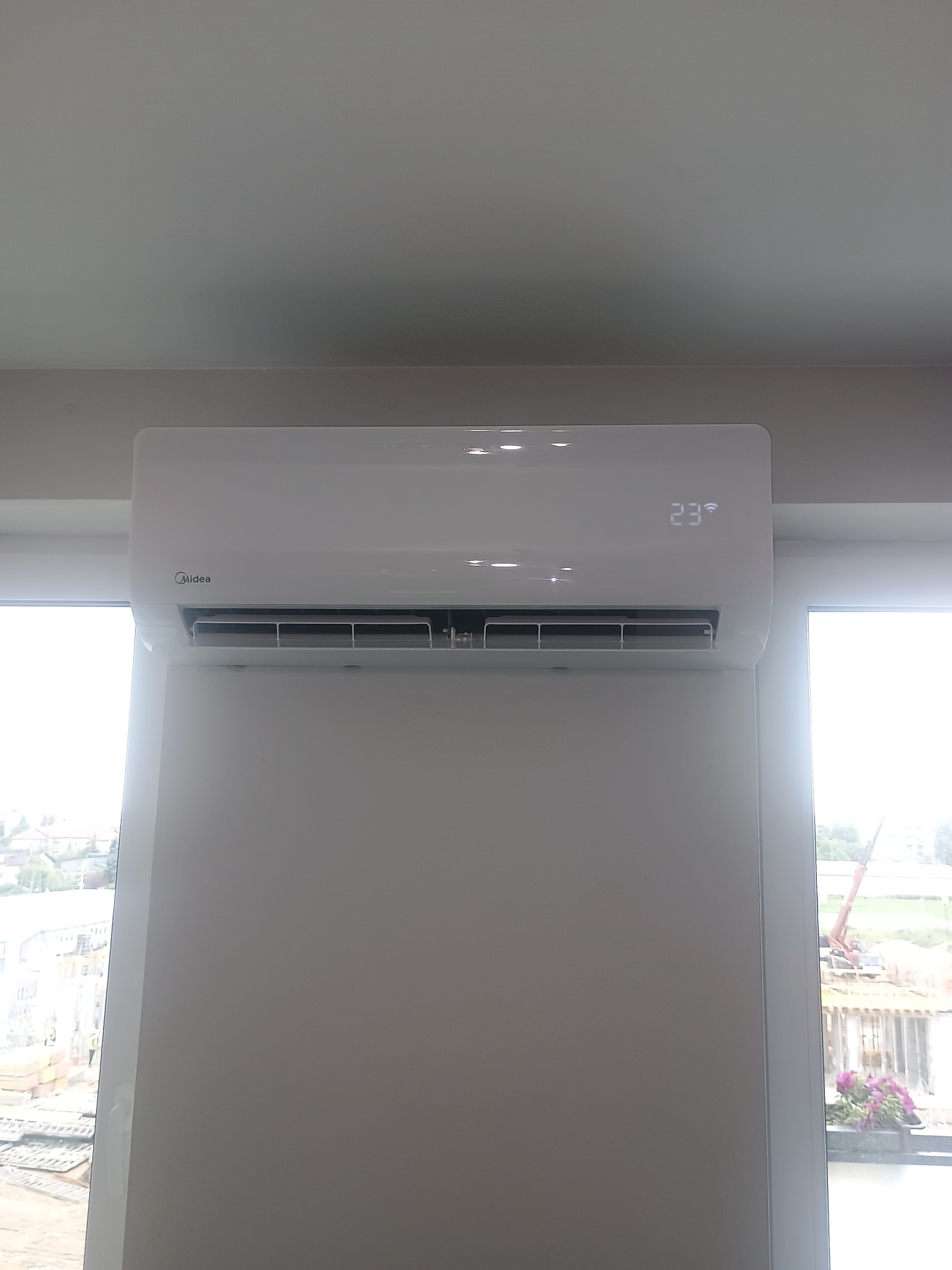 Biały klimatyzator Midea na ścianie, wyświetlający temperaturę 23 stopnie, z widokiem na budowę za oknem. Nowoczesny design.