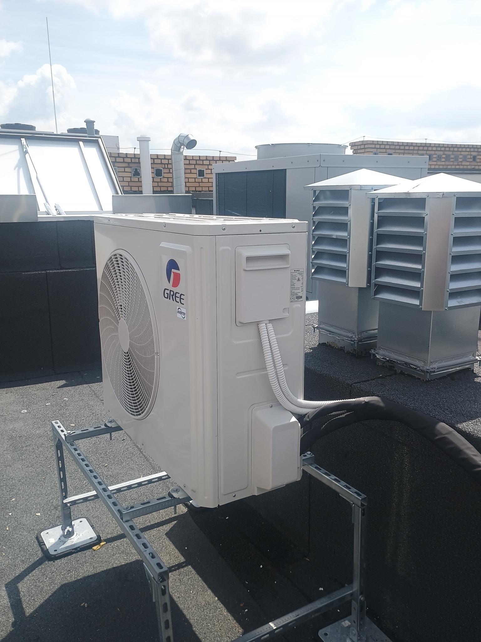 Biała jednostka zewnętrzna klimatyzacji Gree na dachu budynku, zamontowana na metalowej konstrukcji wsporczej. Widoczne przewody i inne elementy instalacji.