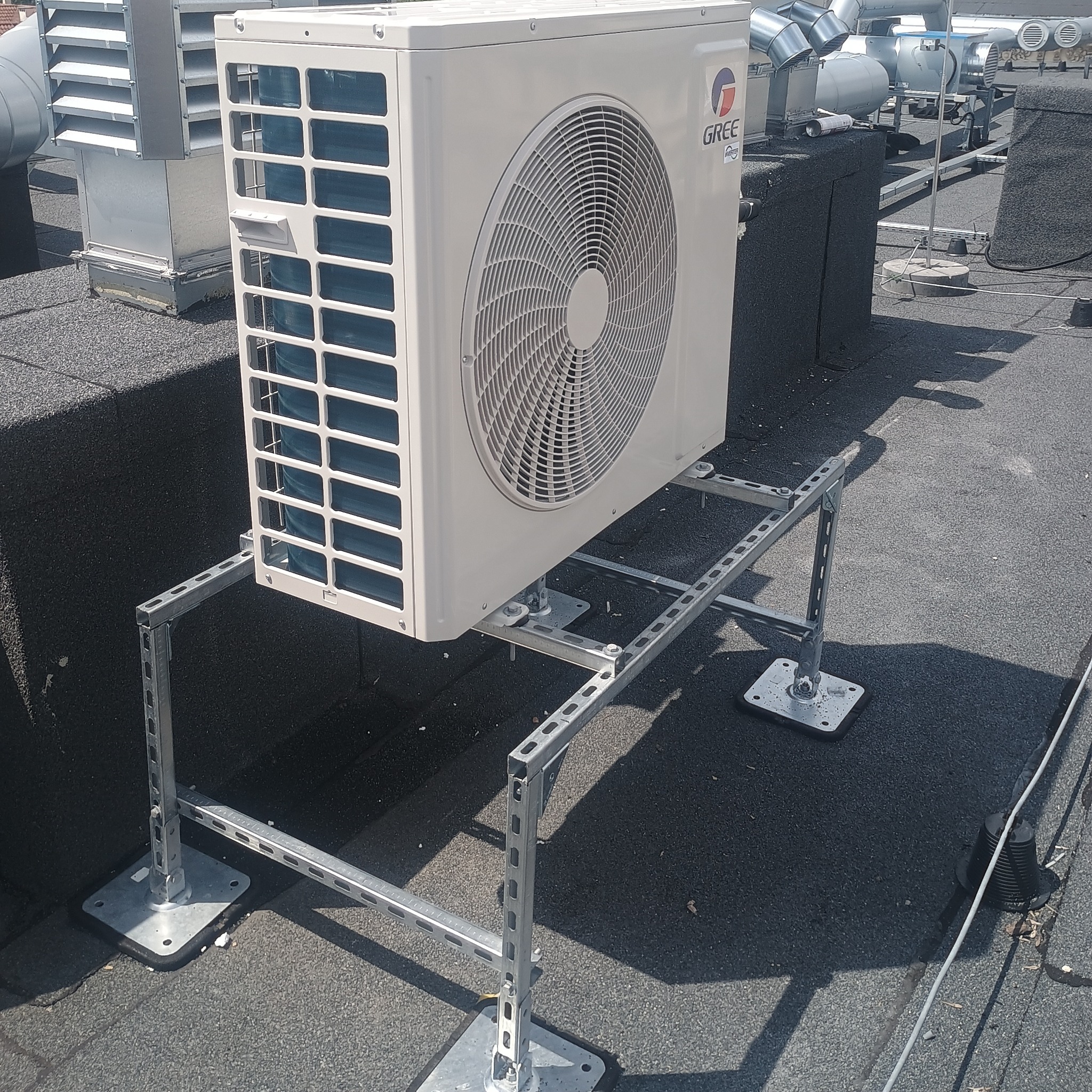 Klimatyzator marki Gree zamontowany na dachu budynku na metalowej konstrukcji wsporczej. Widoczne inne elementy instalacji wentylacyjnej na dachu.