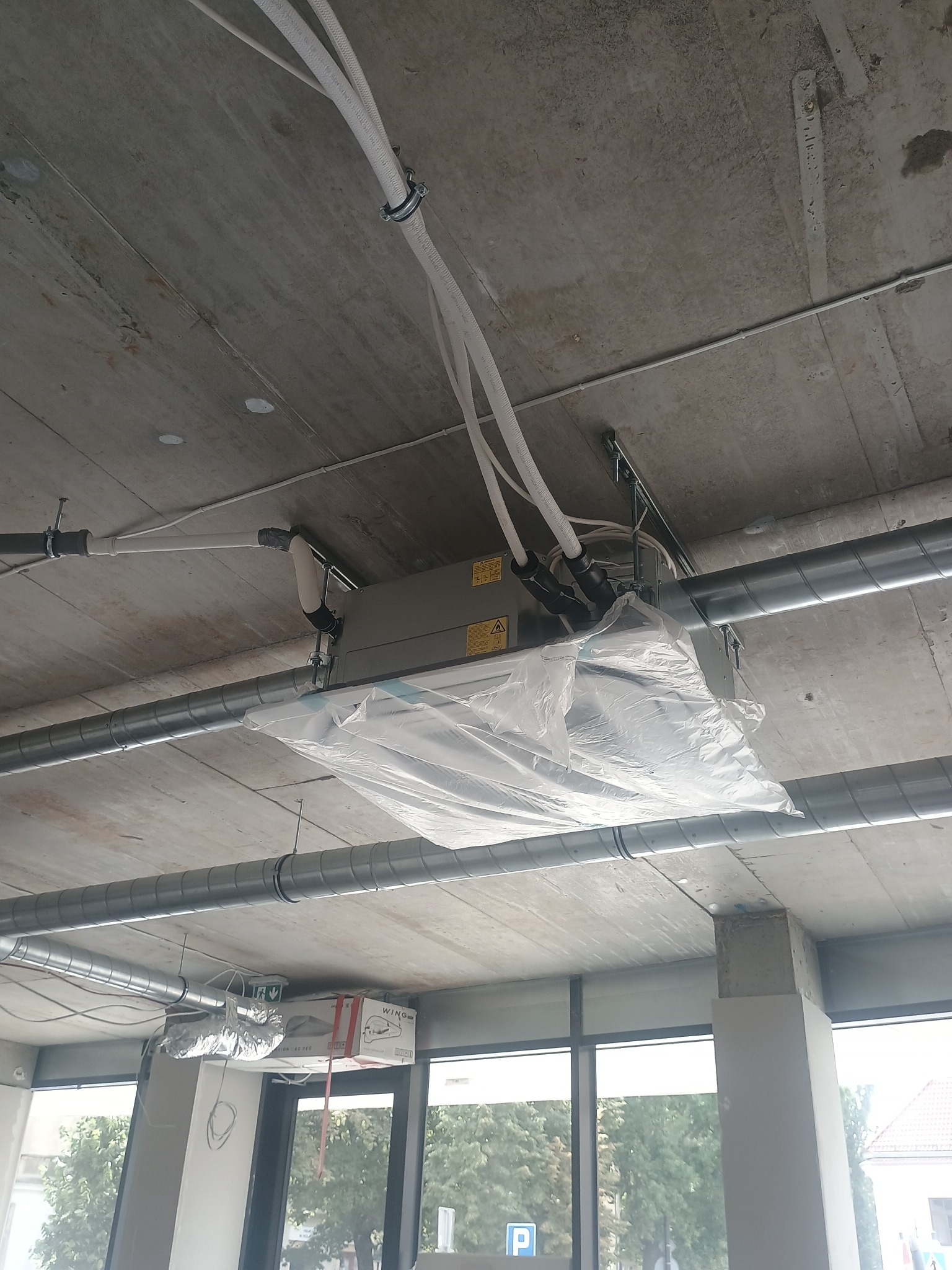 Montaż klimatyzacji kasetonowej pod sufitem w surowym wnętrzu. Widoczne rury wentylacyjne, kable i folia ochronna na urządzeniu. Ujęcie z dołu, surowy beton.