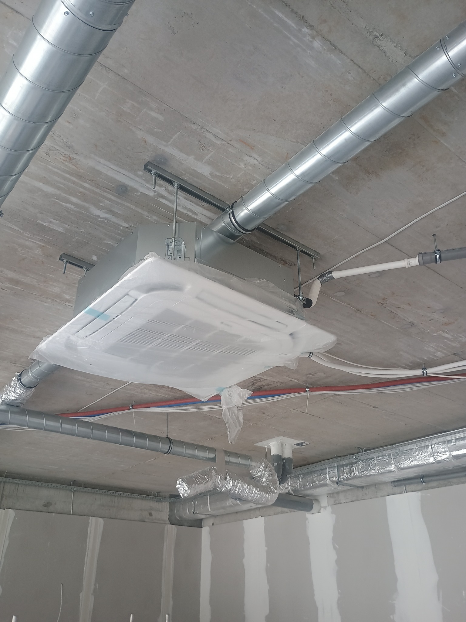 Kasetonowa jednostka klimatyzacji pod sufitem w folii ochronnej, widoczne rury wentylacyjne i instalacje. Surowy betonowy sufit i ściany w trakcie wykończenia.