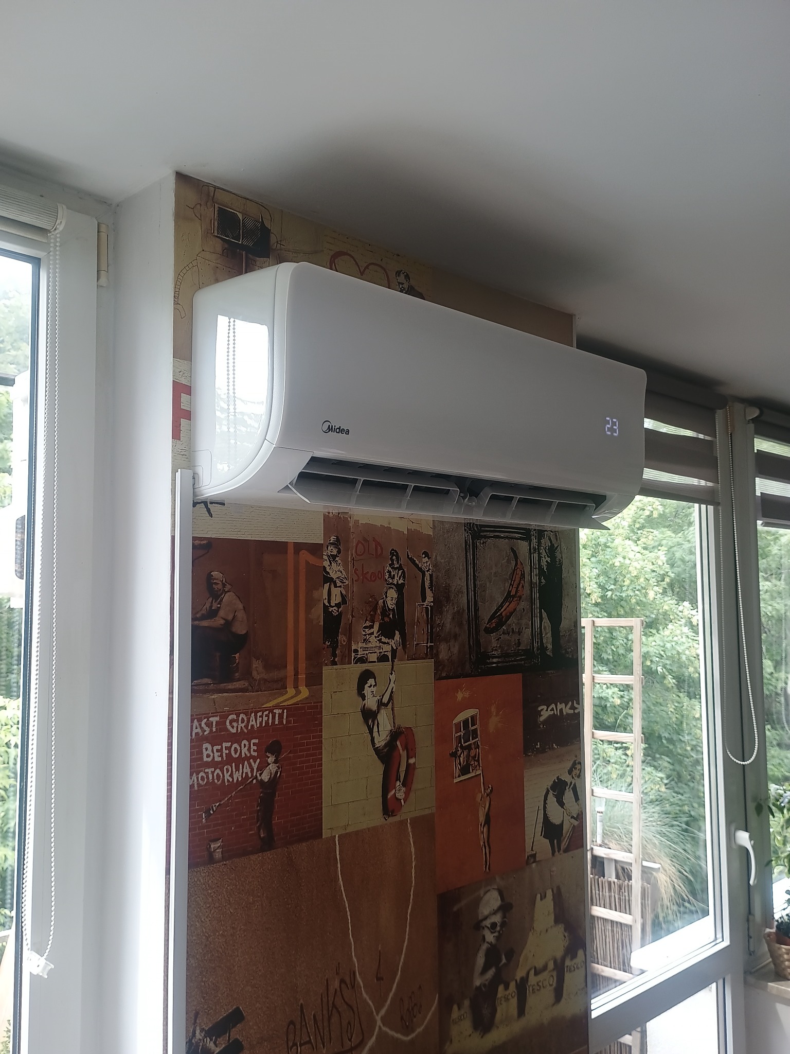 Biały klimatyzator Midea na ścianie z tapetą w stylu graffiti, widoczny fragment okna z zielenią na zewnątrz. Wyświetlacz klimatyzatora pokazuje temperaturę 23 stopnie.