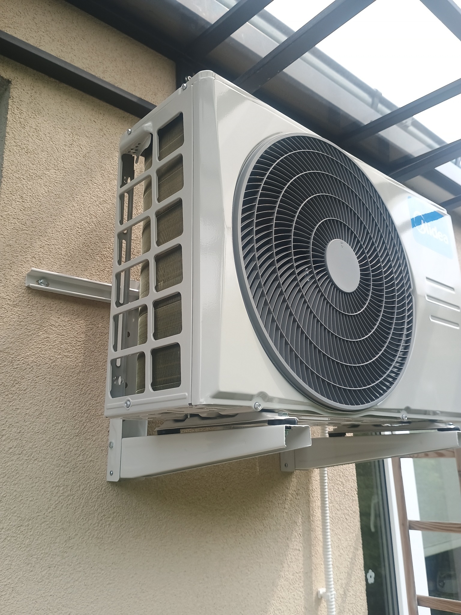 Zamontowana jednostka zewnętrzna klimatyzacji Midea na elewacji budynku, widoczna z boku, z elementami konstrukcji wsporczej i fragmentem zadaszenia.