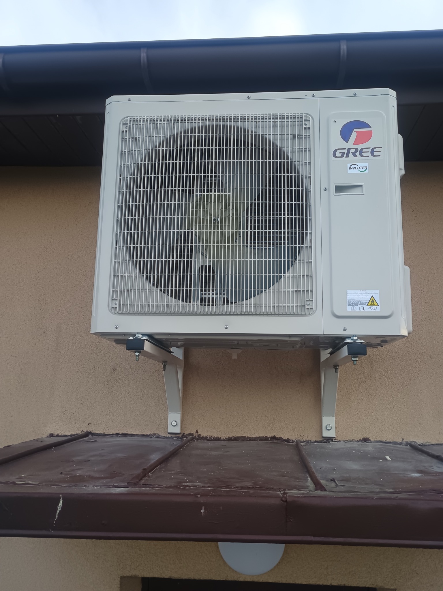 Zewnętrzna jednostka klimatyzacji GREE zamontowana na ścianie budynku, widoczna z bliska. Szara obudowa, metalowa kratka wentylatora i logo producenta.