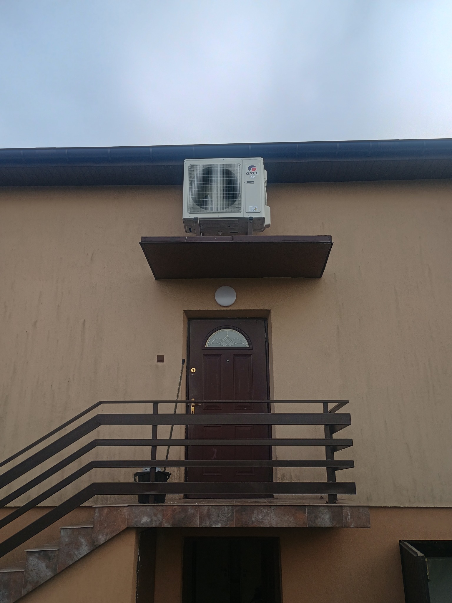 Biała jednostka zewnętrzna klimatyzacji marki Gree zamontowana nad wejściem do budynku z brązowymi drzwiami i schodami z balustradą. Widok z dołu.