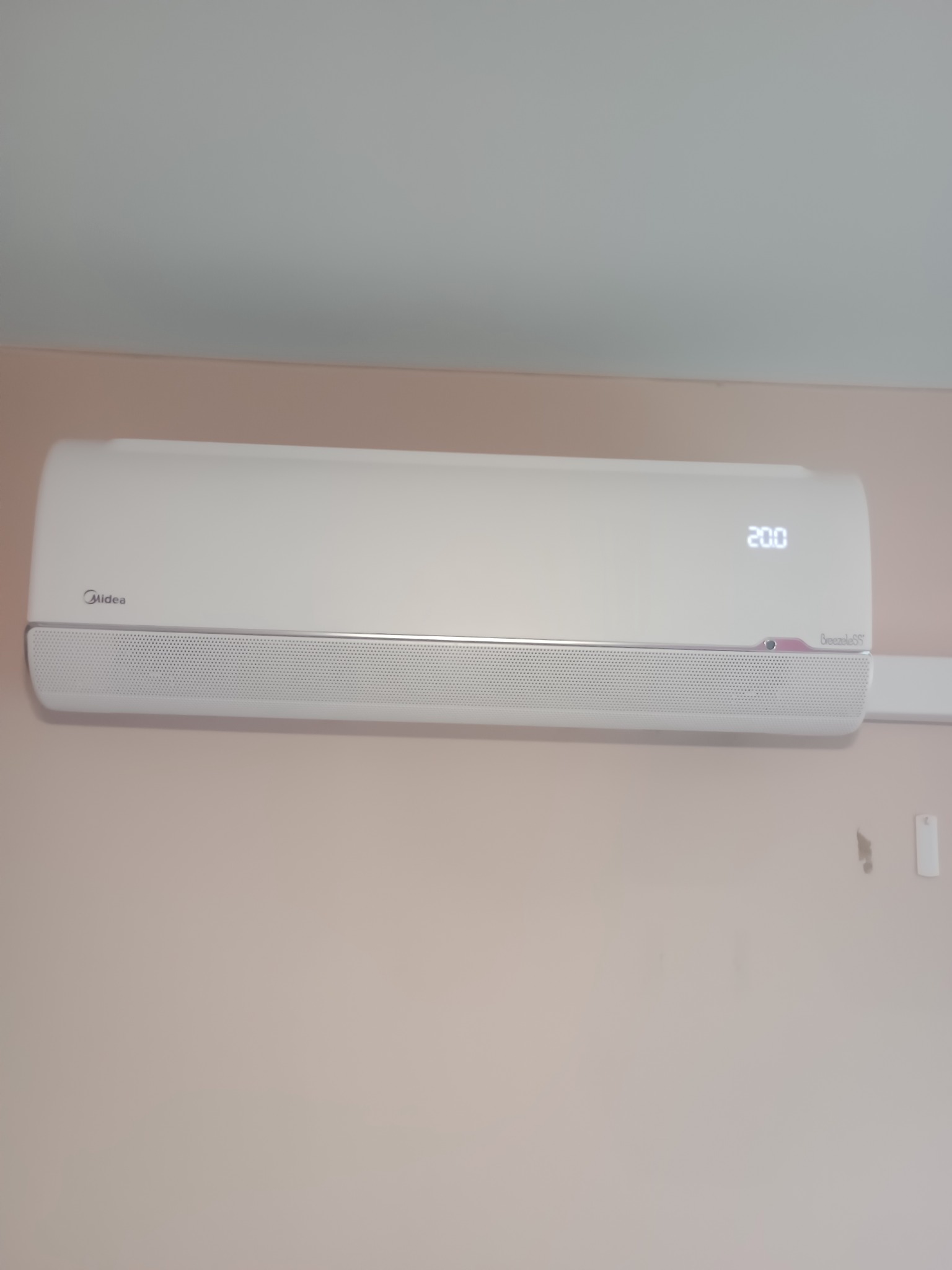 Biały klimatyzator Midea Breezeless z wyświetlaczem 20.0 zamontowany na jasnoróżowej ścianie. Widoczny fragment sufitu i element instalacji elektrycznej.