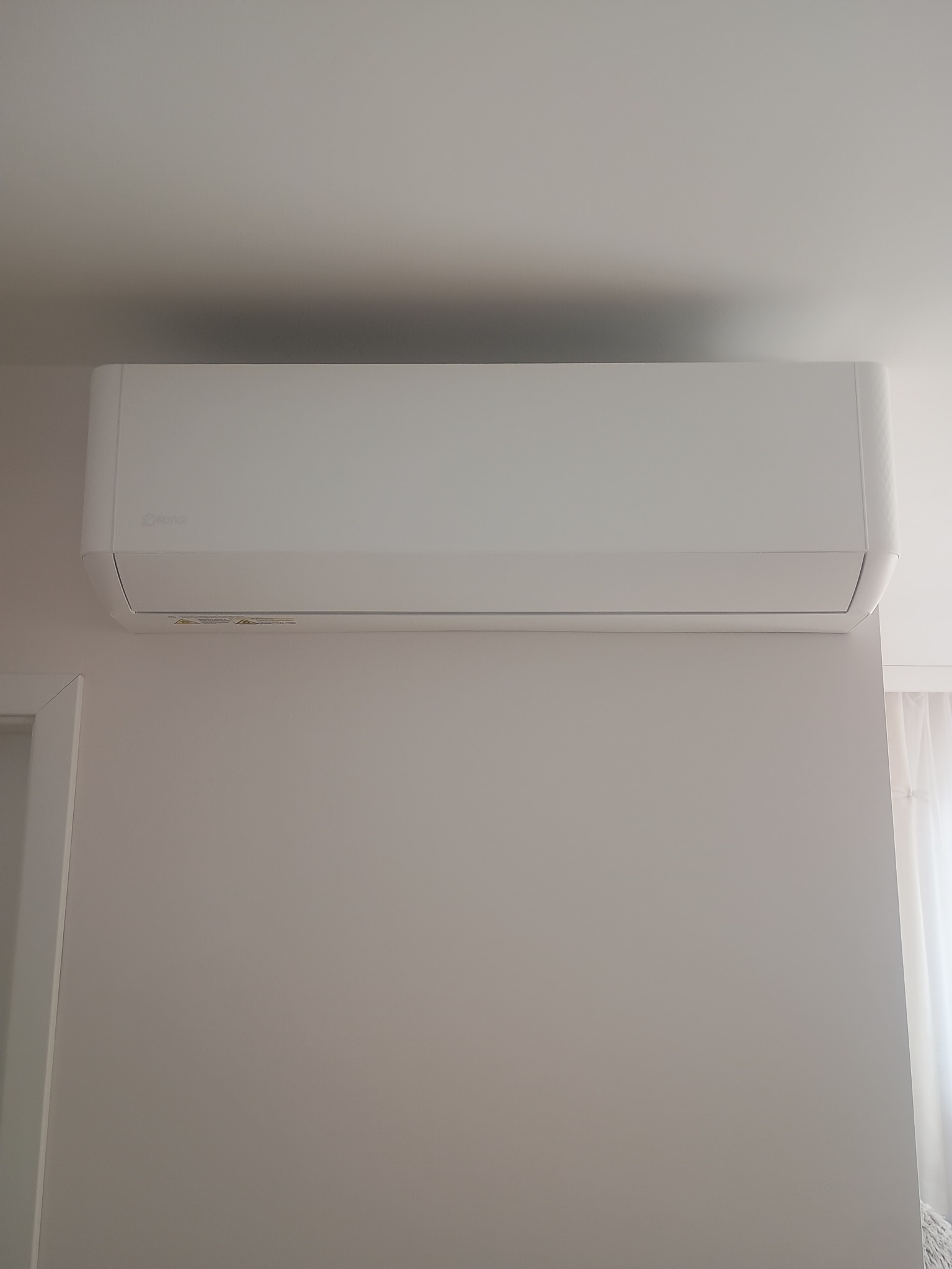 Biała, minimalistyczna klimatyzacja ścienna marki Konsel, zamontowana w jasnym pomieszczeniu. Widoczny fragment futryny drzwi i zasłony w tle.