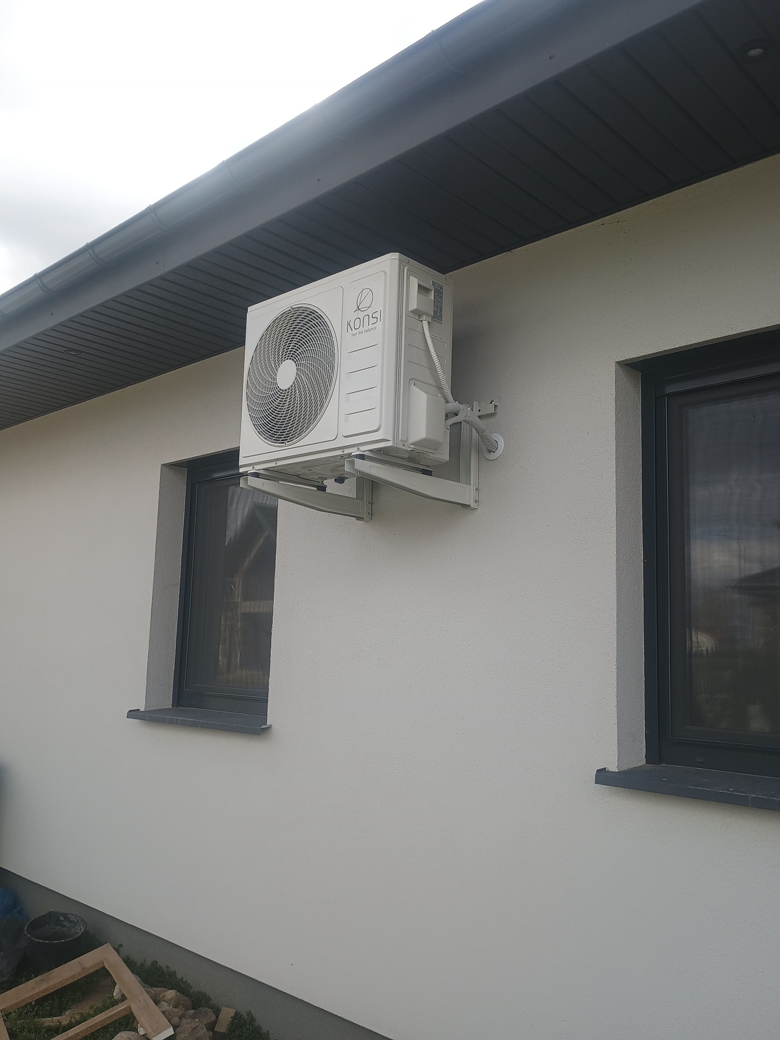 Biała jednostka zewnętrzna klimatyzacji marki Konsi zamontowana na jasnej elewacji domu, widoczna z boku, z szarymi ramami okiennymi i ciemnym podbitką dachową.