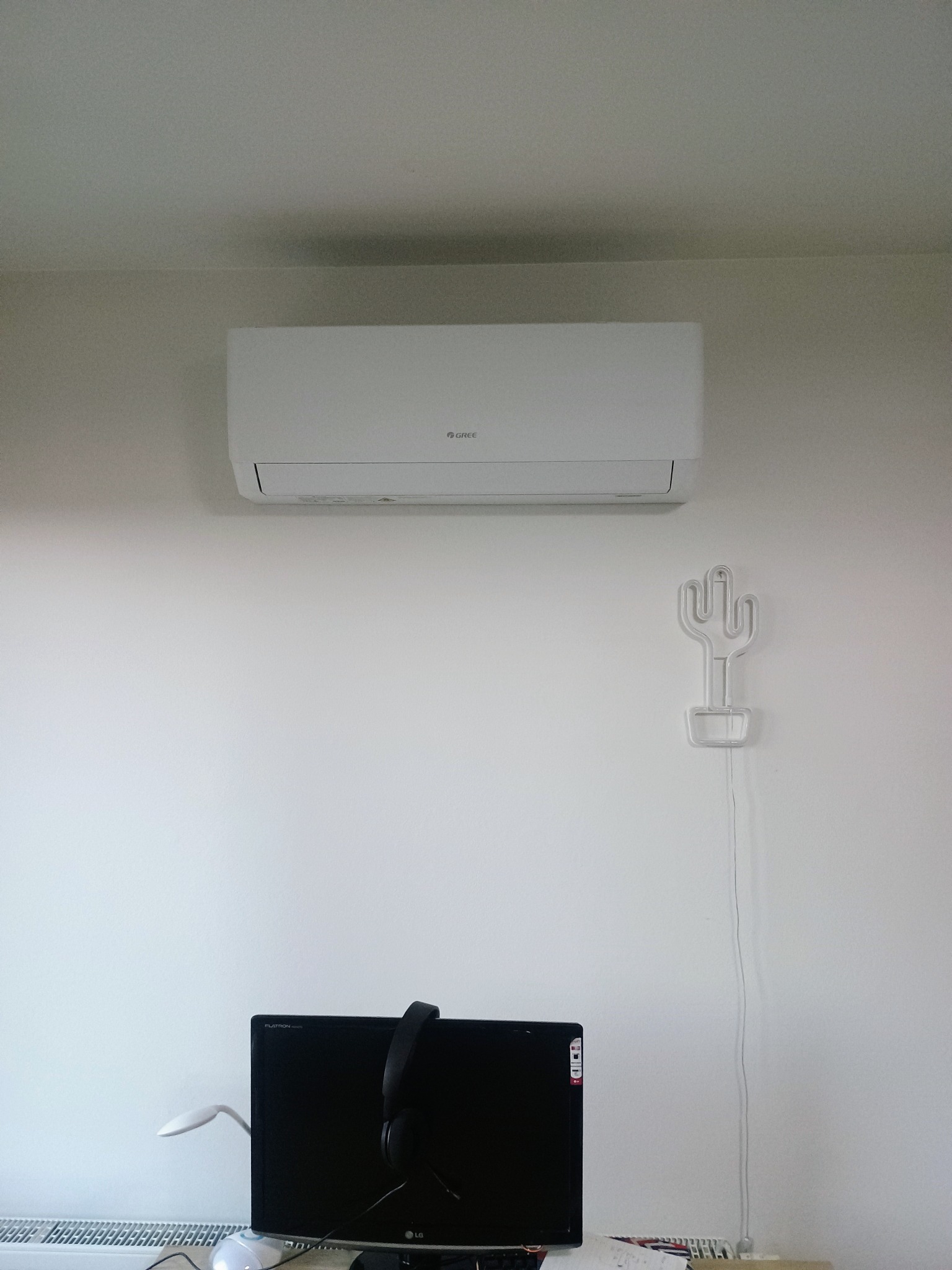 Biały klimatyzator marki Gree zamontowany na ścianie, pod nim neon w kształcie kaktusa i monitor. Widok z dołu, oświetlenie dzienne.