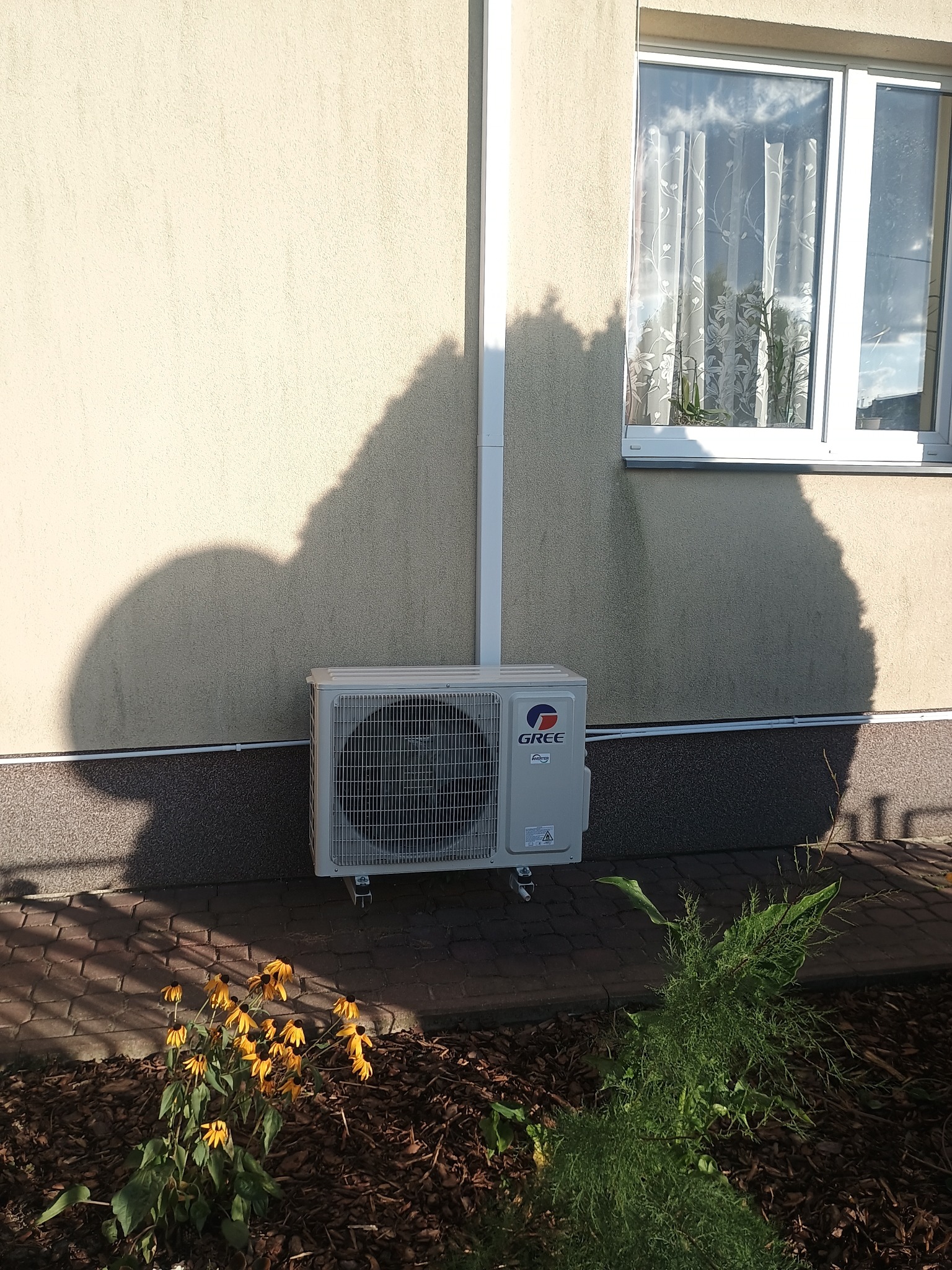 Zewnętrzna jednostka klimatyzacji Gree zamontowana na elewacji budynku, z widocznym oknem i roślinnością w tle. Cień drzewa na ścianie.