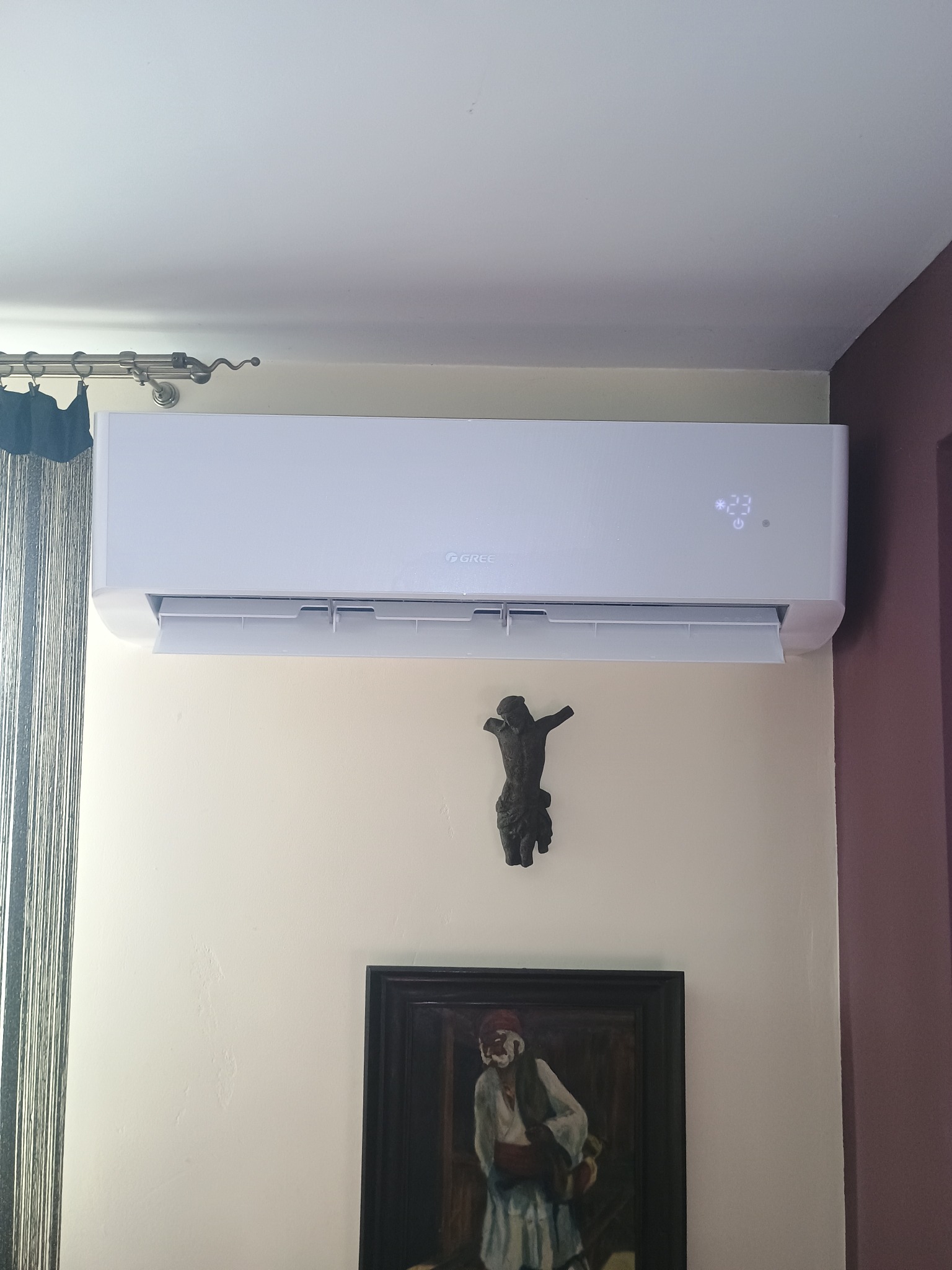 Biały klimatyzator Gree zamontowany na ścianie w pomieszczeniu, pod nim wisi krucyfiks i obraz w czarnej ramie. Widoczny fragment zasłony i ściany w kolorze bordo.