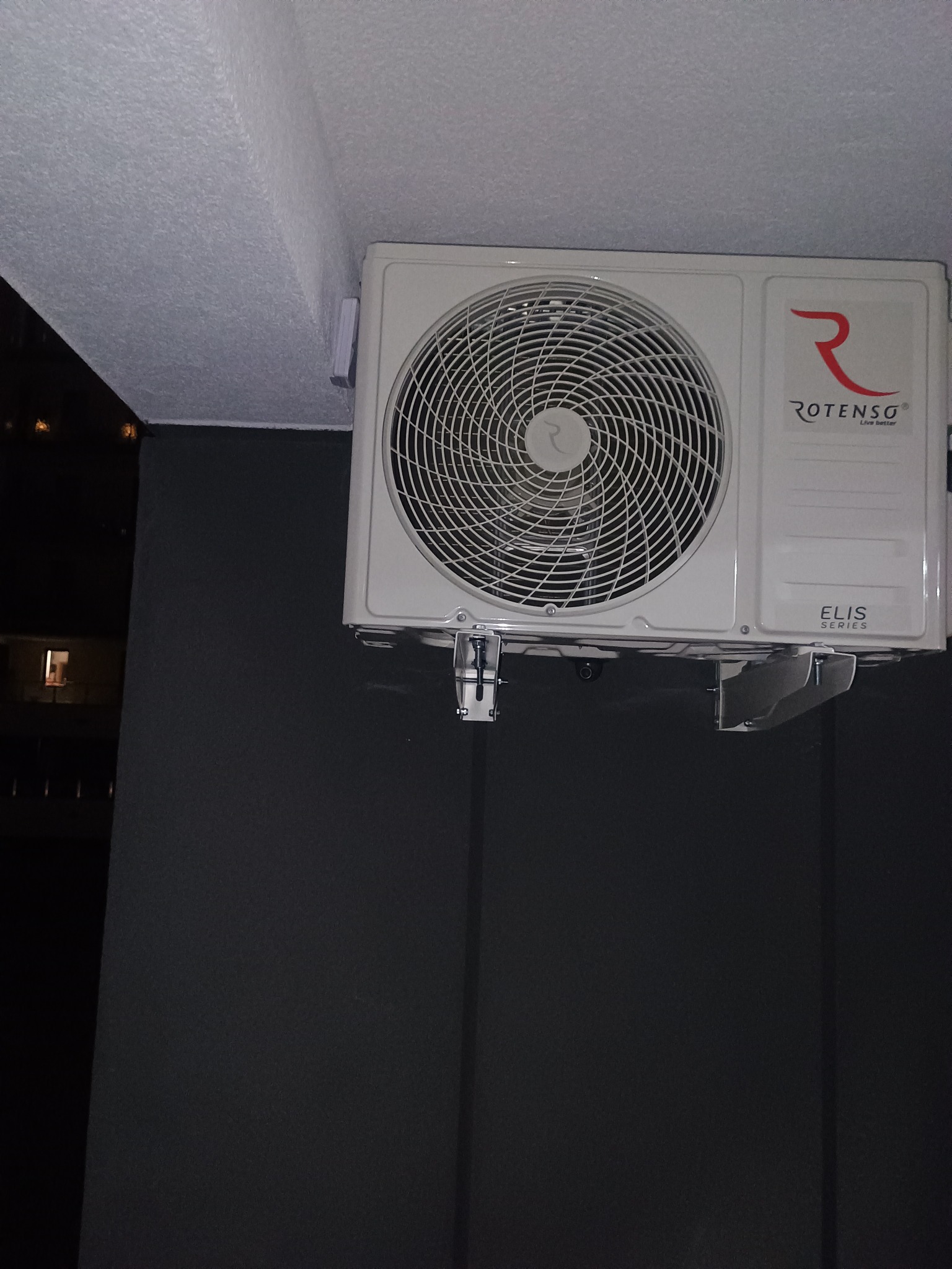 Zamontowany klimatyzator Rotenso Elis Series na ciemnej ścianie balkonu, widoczne mocowania i fragment sufitu. W tle nocny widok z oknem.