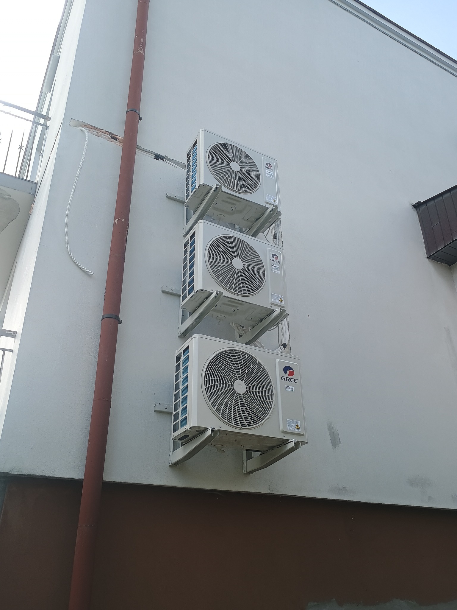 Trzy klimatyzatory Gree zamontowane jeden nad drugim na jasnej ścianie budynku. Widoczna rynna spustowa i fragment balkonu. Ujęcie z dołu, podkreślające pionowy układ urządzeń.