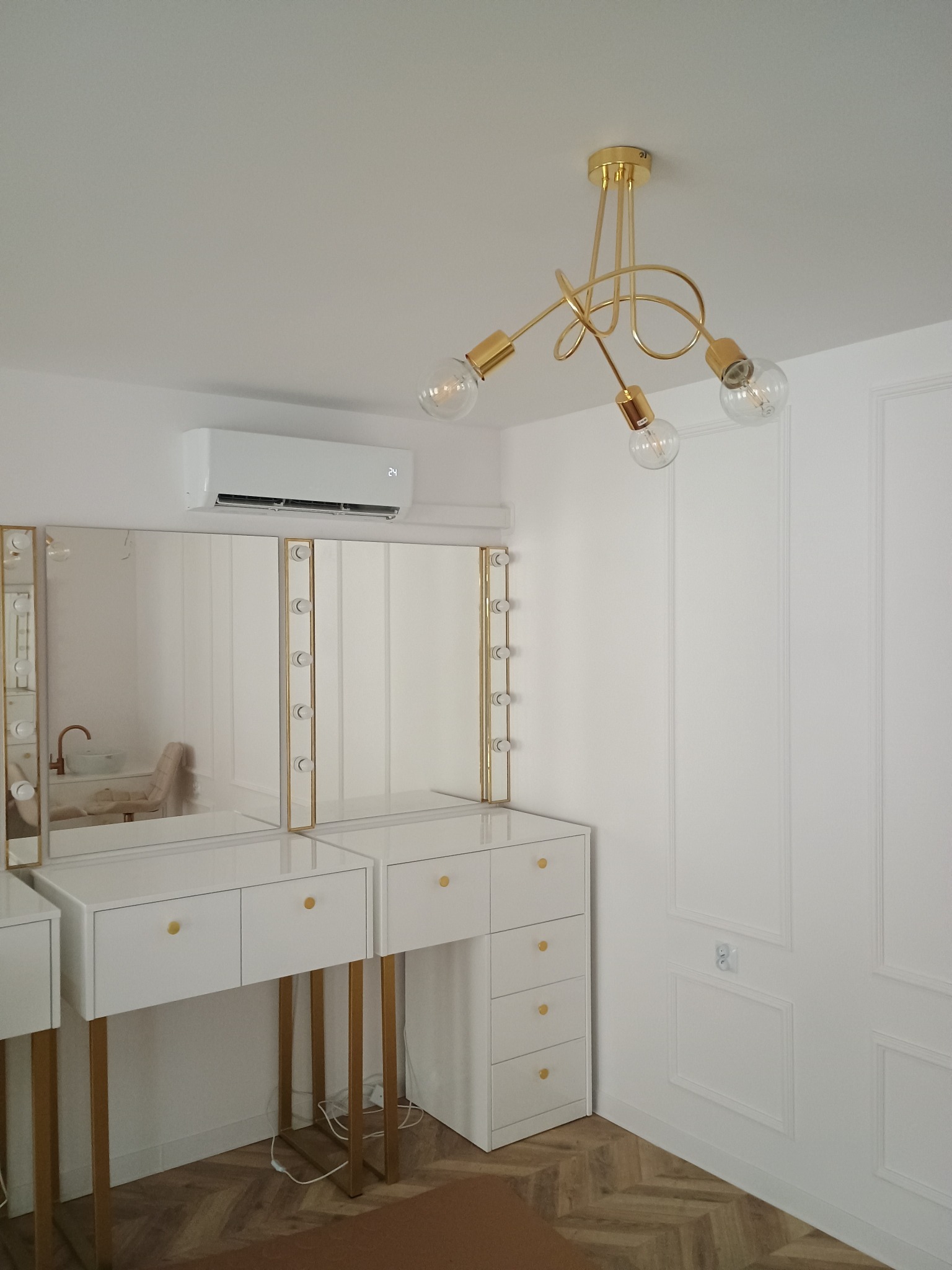 Biała klimatyzacja ścienna nad toaletką z lustrami i oświetleniem w nowoczesnym wnętrzu. Złoty żyrandol dodaje elegancji. Widoczny parkiet i dekoracyjne panele ścienne.