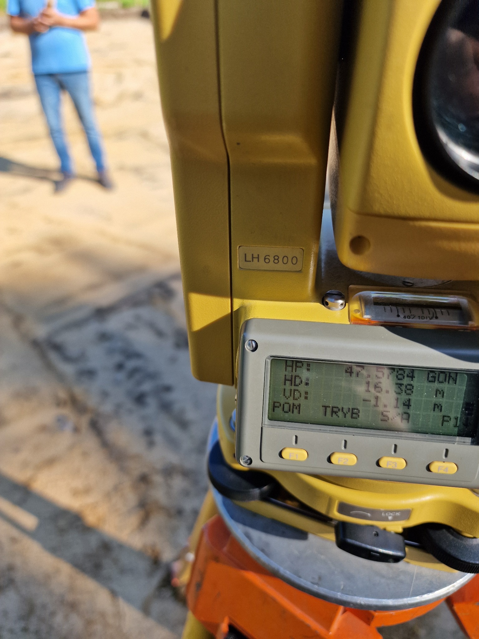 Żółty teodolit z wyświetlaczem LCD pokazującym dane pomiarowe, ustawiony na pomarańczowym statywie, w tle rozmyta sylwetka osoby w niebieskiej koszulce.